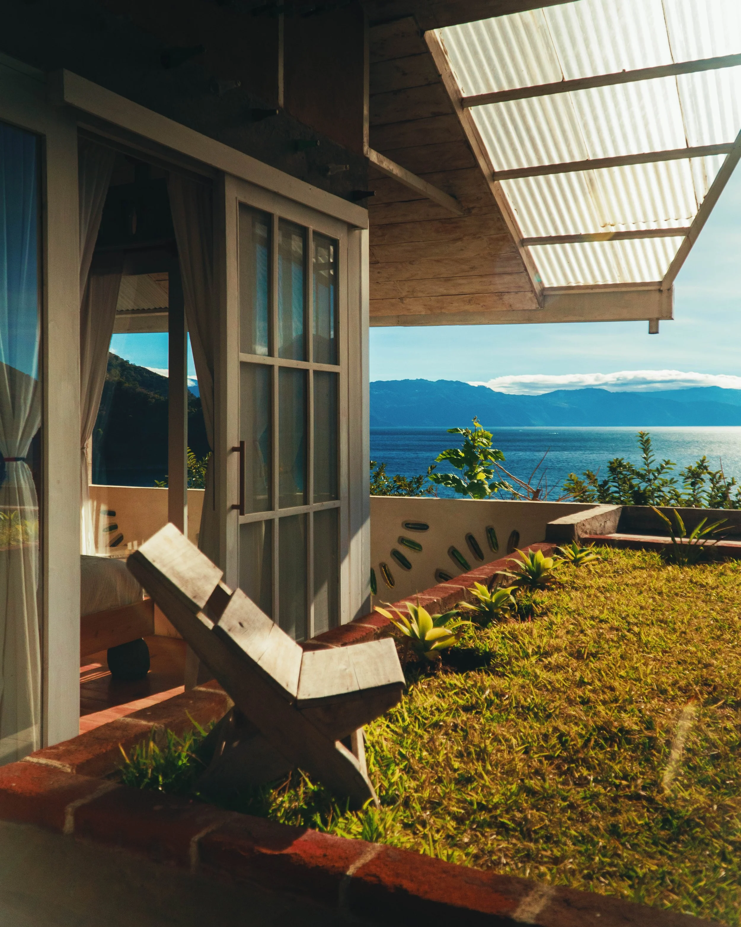 Terraza o balcón con vista a un mar, con plantas en el borde y una cabaña de madera con estructura de vidrio y cortinas, y un sillón de madera en primer plano, en una zona montañosa con cielo azul y nubes.