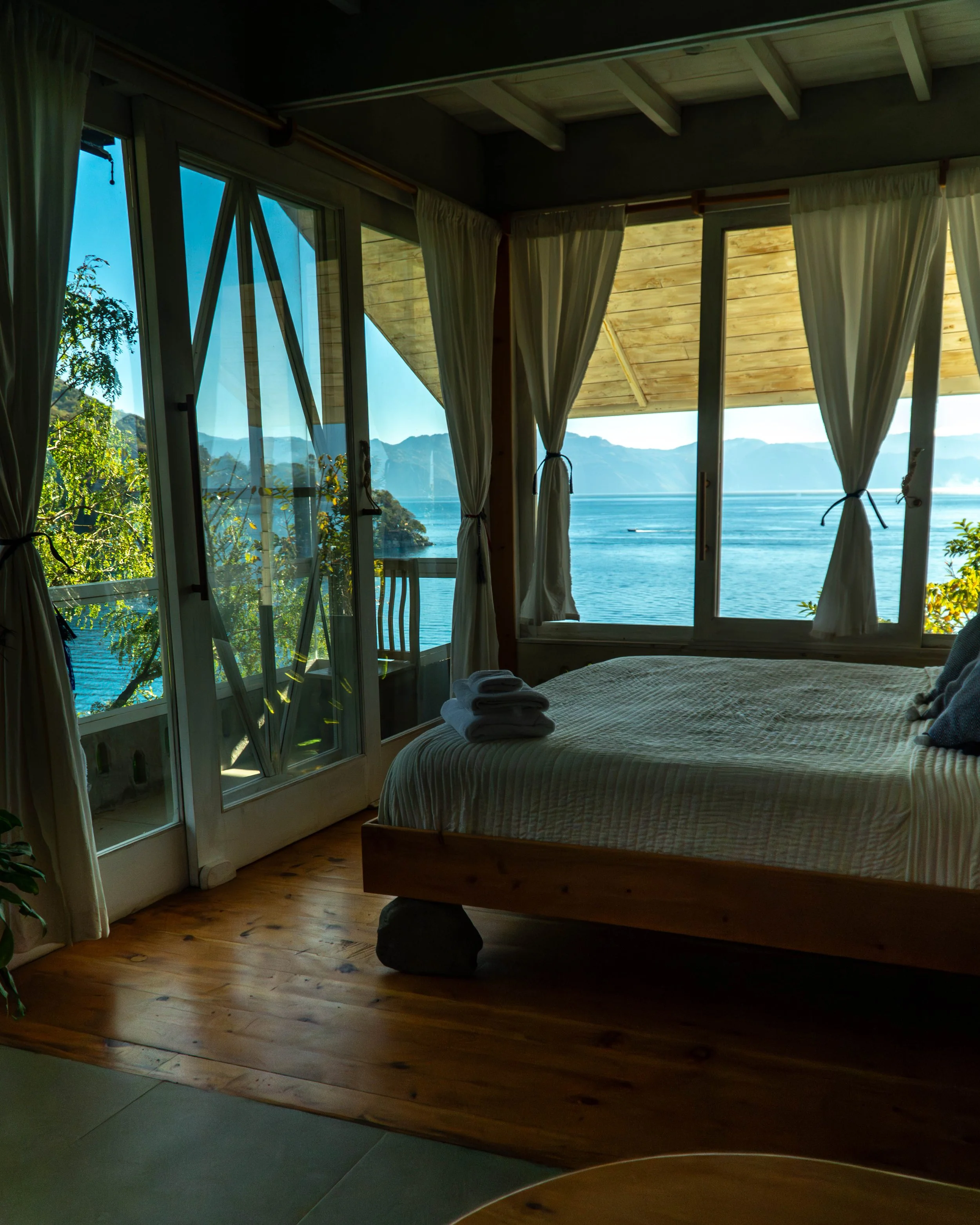 Habitación con vista al lago y montañas, con grandes ventanas y cortinas blancas, cama con ropa blanca y toallas dobladas.
