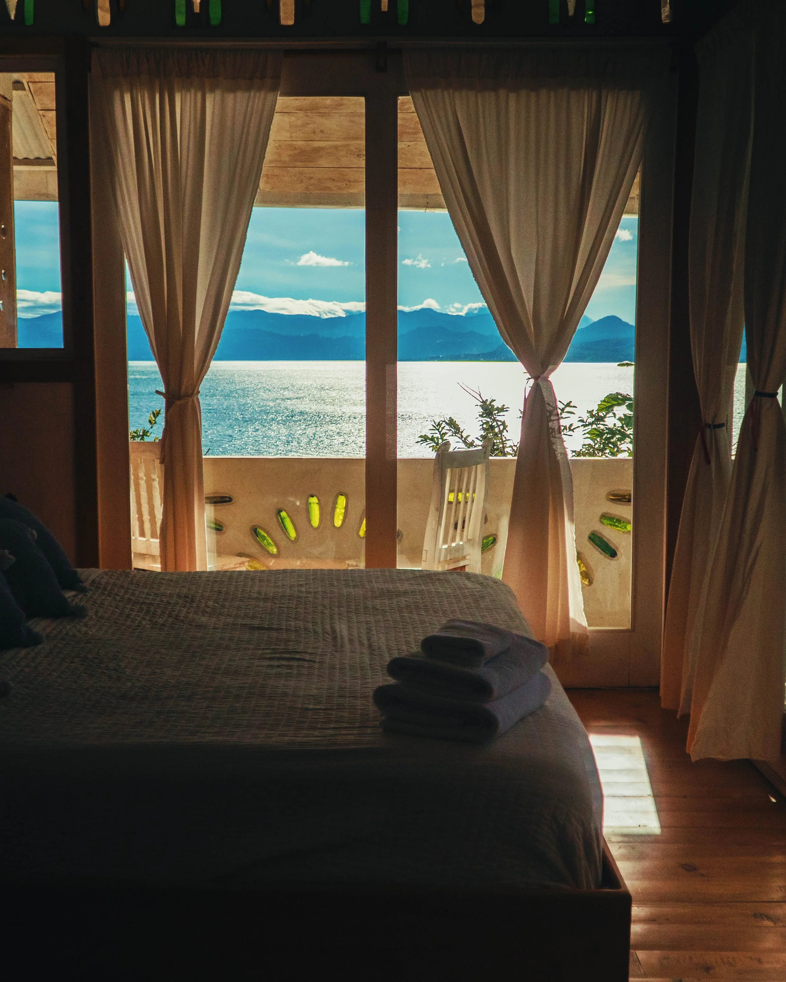 Vista desde una habitación que muestra una ventana con cortinas blancas, un balcón con sillas blancas y un paisaje de montañas y agua al fondo bajo un cielo con algunas nubes.