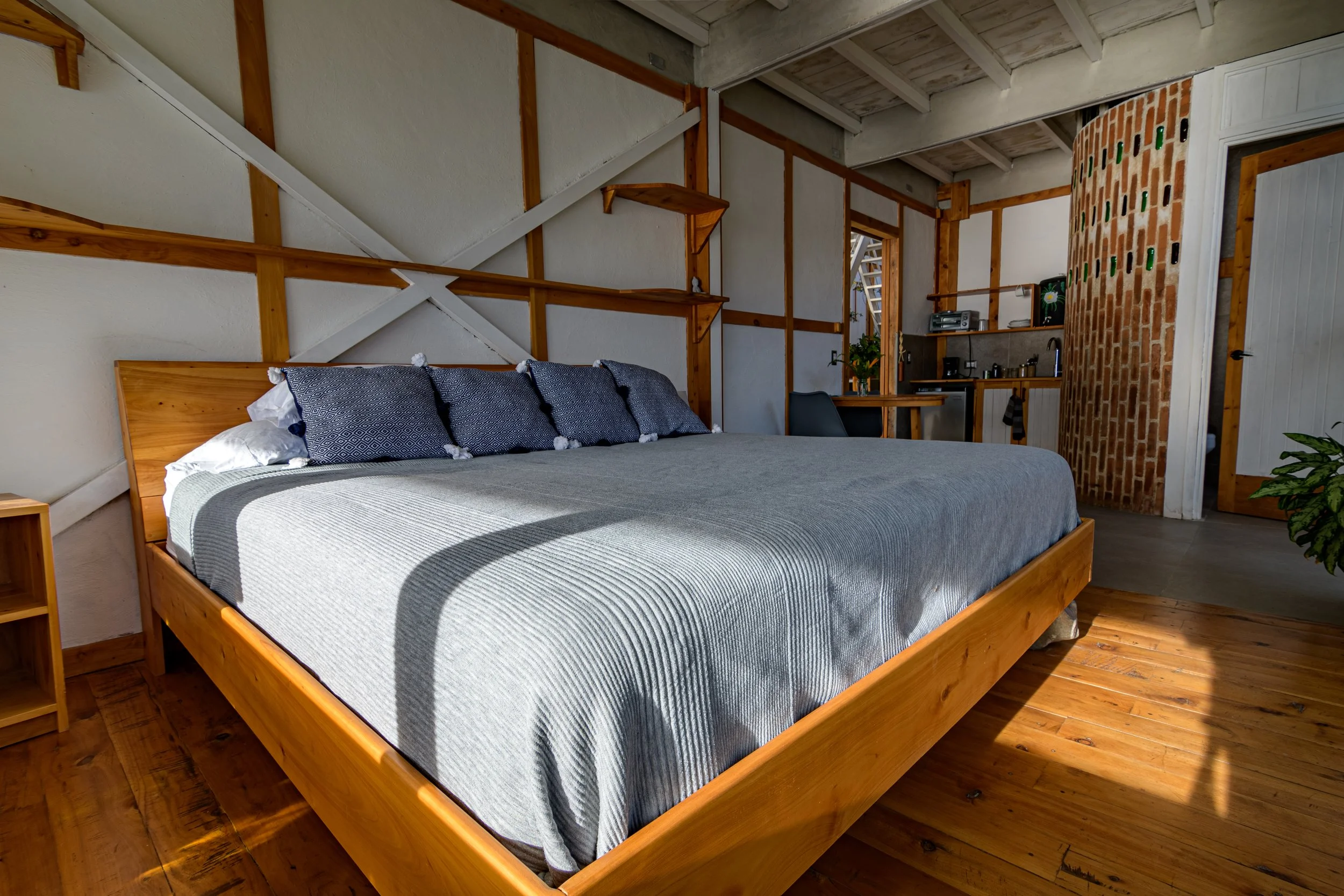 Una habitación con una cama de madera con sábanas blancas y almohadas azules. La pared del fondo tiene un diseño con madera y estanterías. Hay una pequeña área de cocina con electrodomésticos y una mesa con flores. Puertas y ventanas permiten la entr