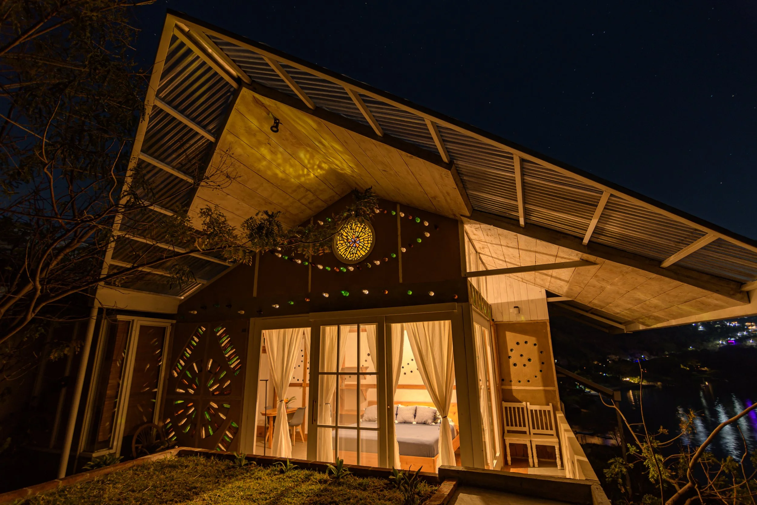 Vista nocturna de una casa moderna con ventanas de vidrio, decorada con luces de colores, bajo un cielo estrellado.