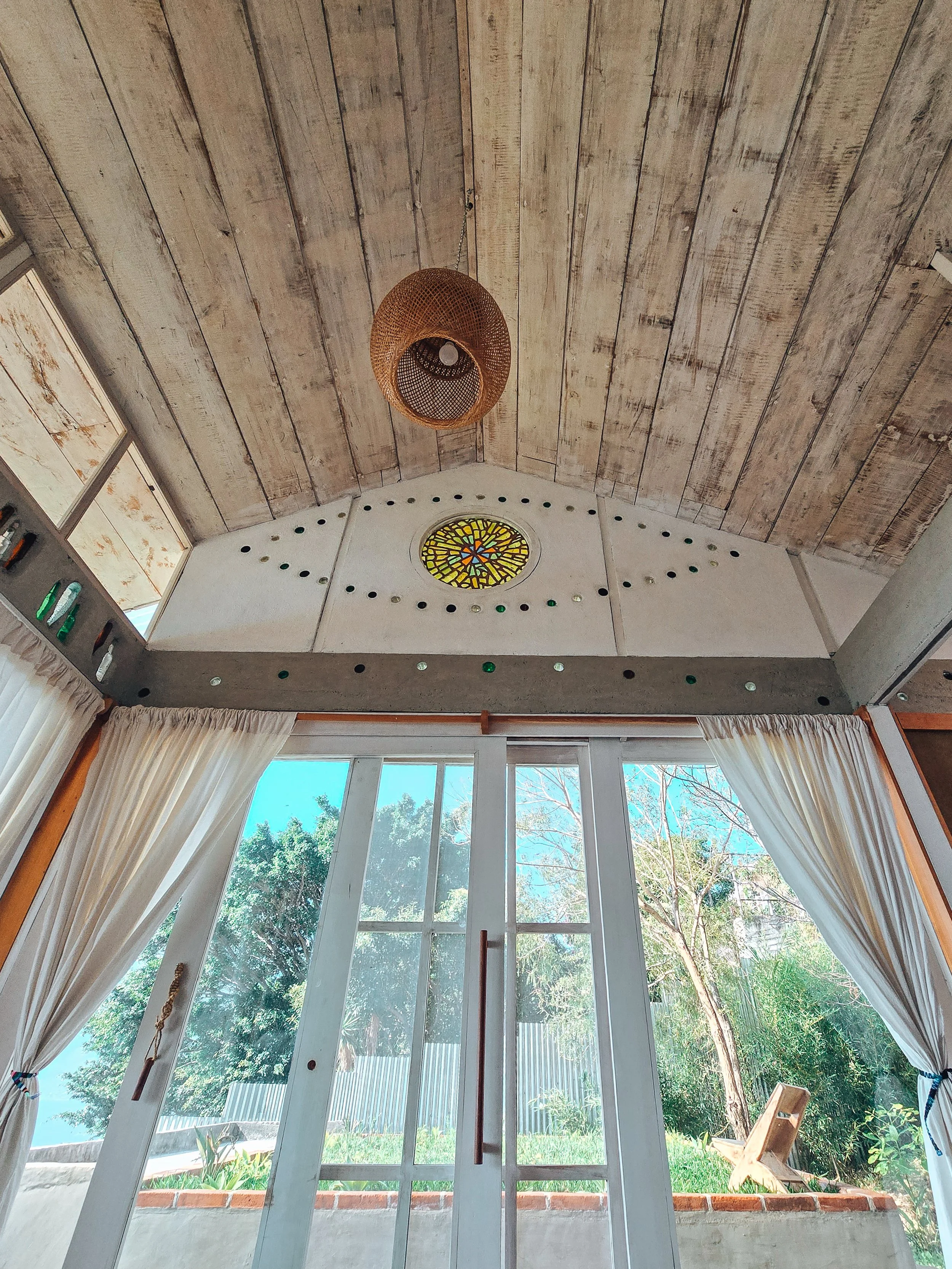 Persiana y ventanas de cristal en una puerta que da al jardín con árboles y césped, con cortinas blancas.
