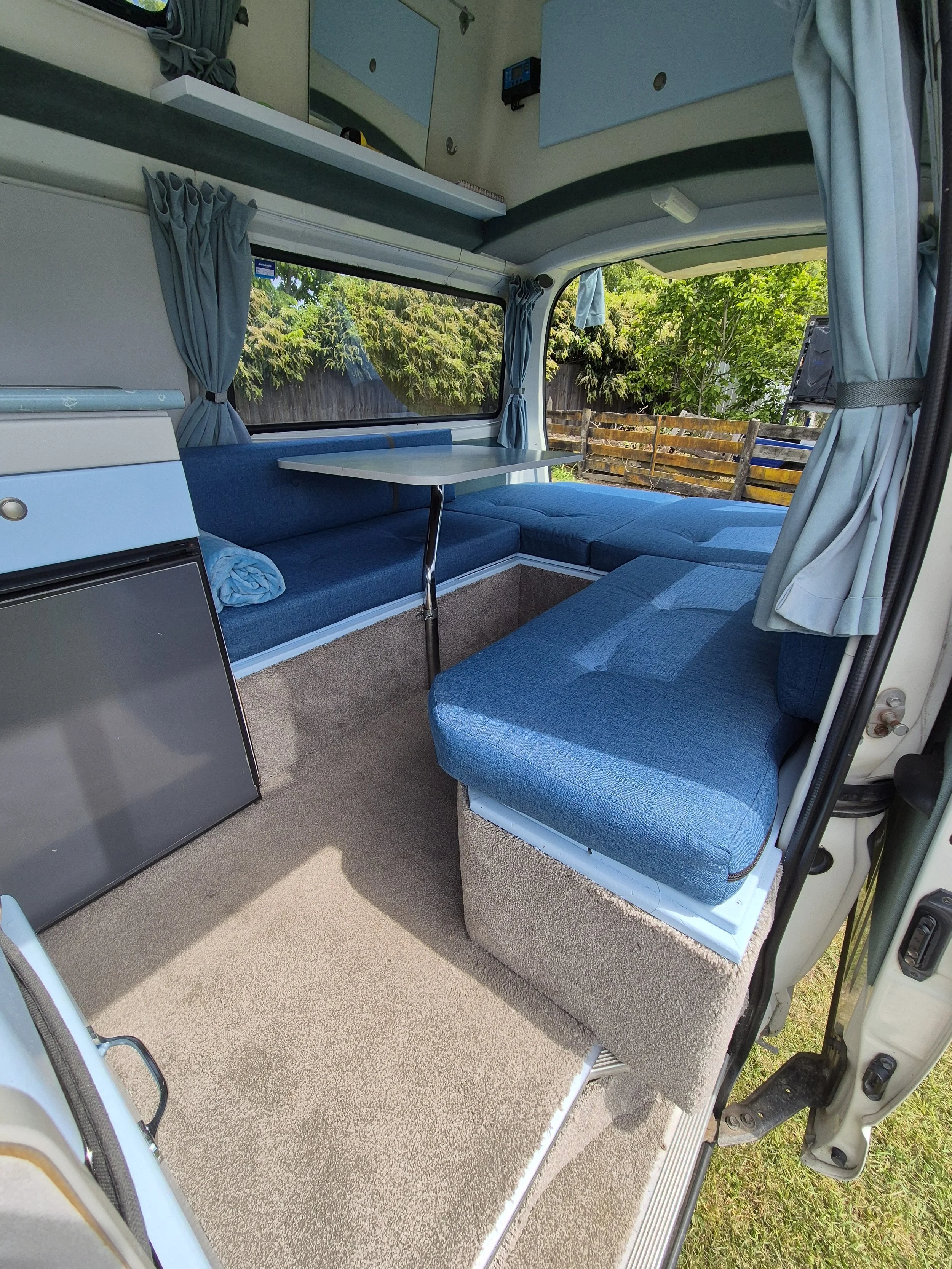 Interior of a camper with a blue cushioned bench, a small white table, windows showing a lake and mountains, kitchen sink, and stove.