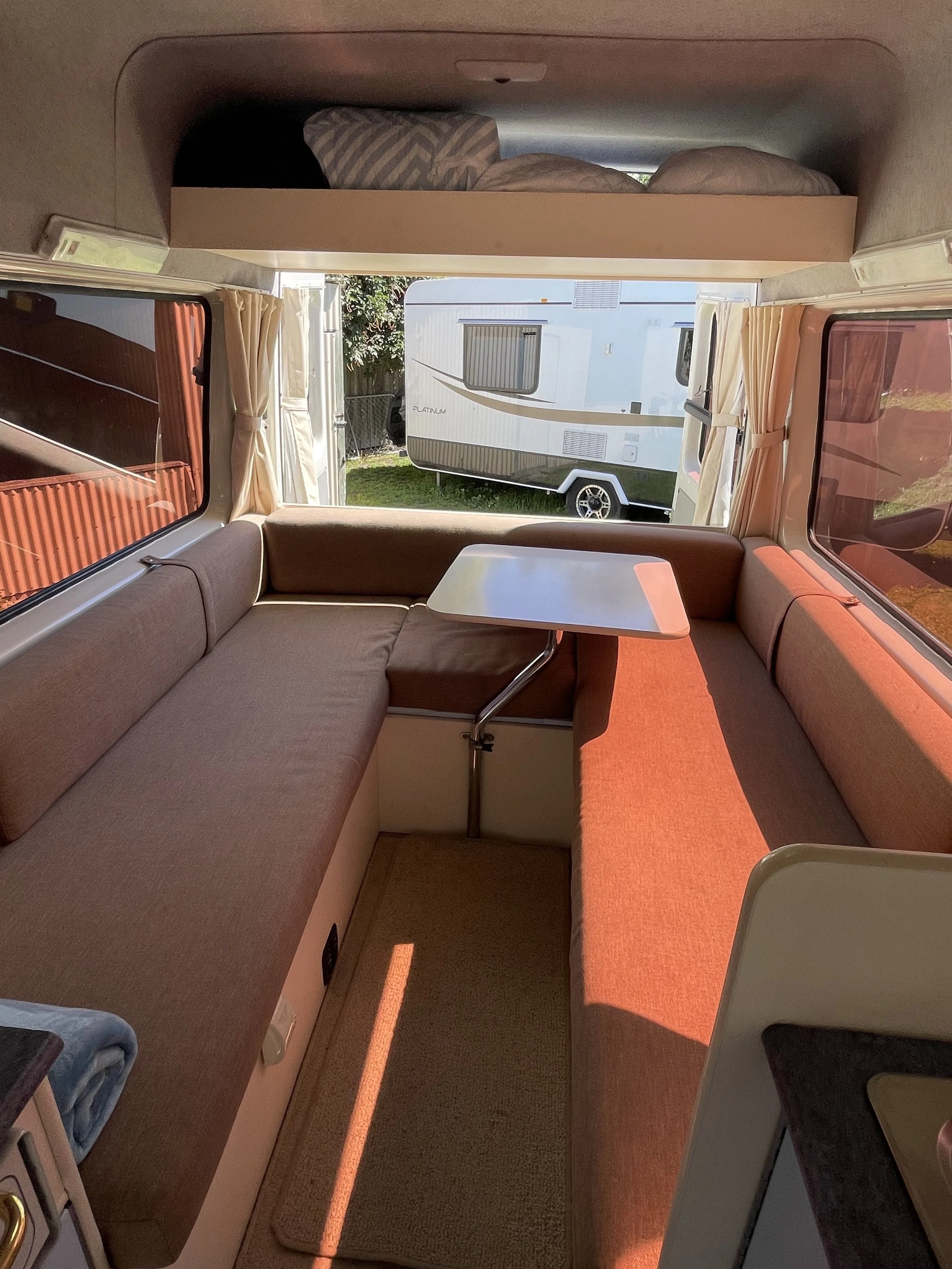 Interior of a camper with a blue cushioned bench, a small white table, windows showing a lake and mountains, kitchen sink, and stove.