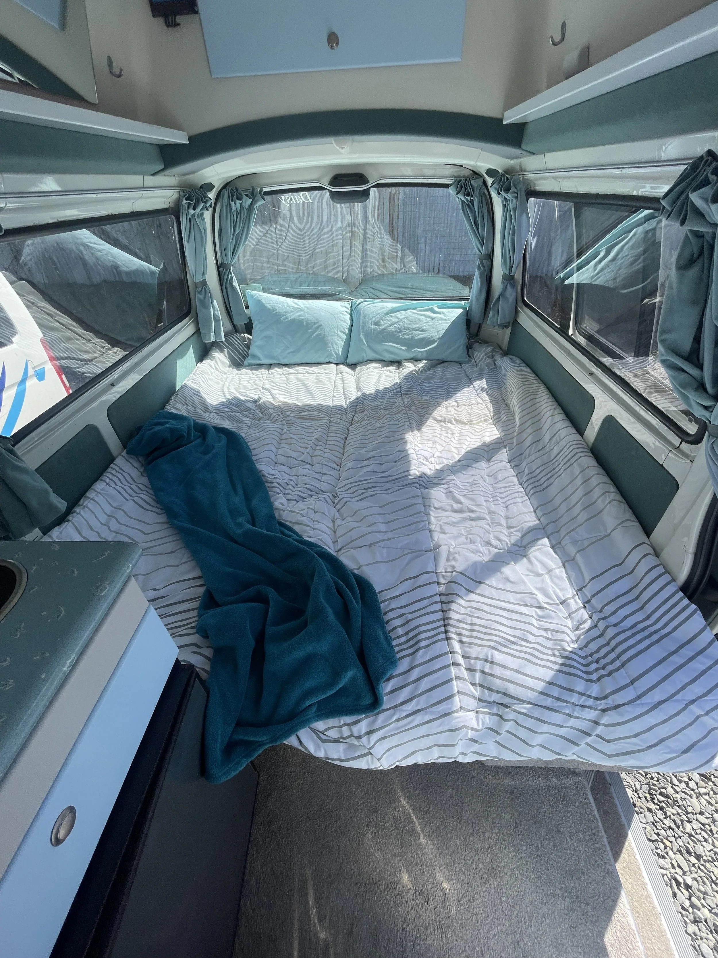 Bedroom inside a camper with a large window showing a scenic view of mountains, lake, and trees, with beige curtains and a blue bed covering.