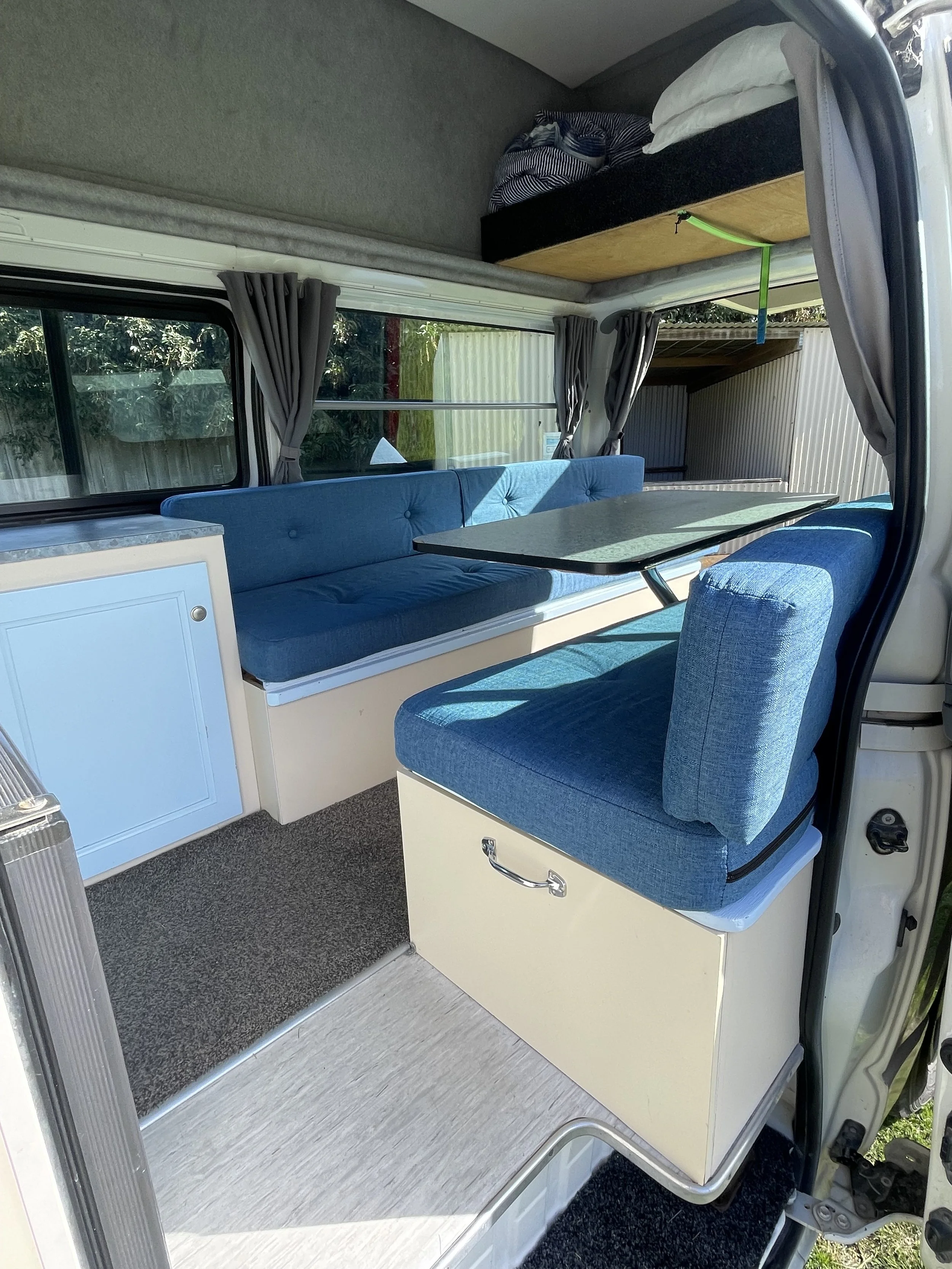 Interior of a camper with a blue cushioned bench, a small white table, windows showing a lake and mountains, kitchen sink, and stove.