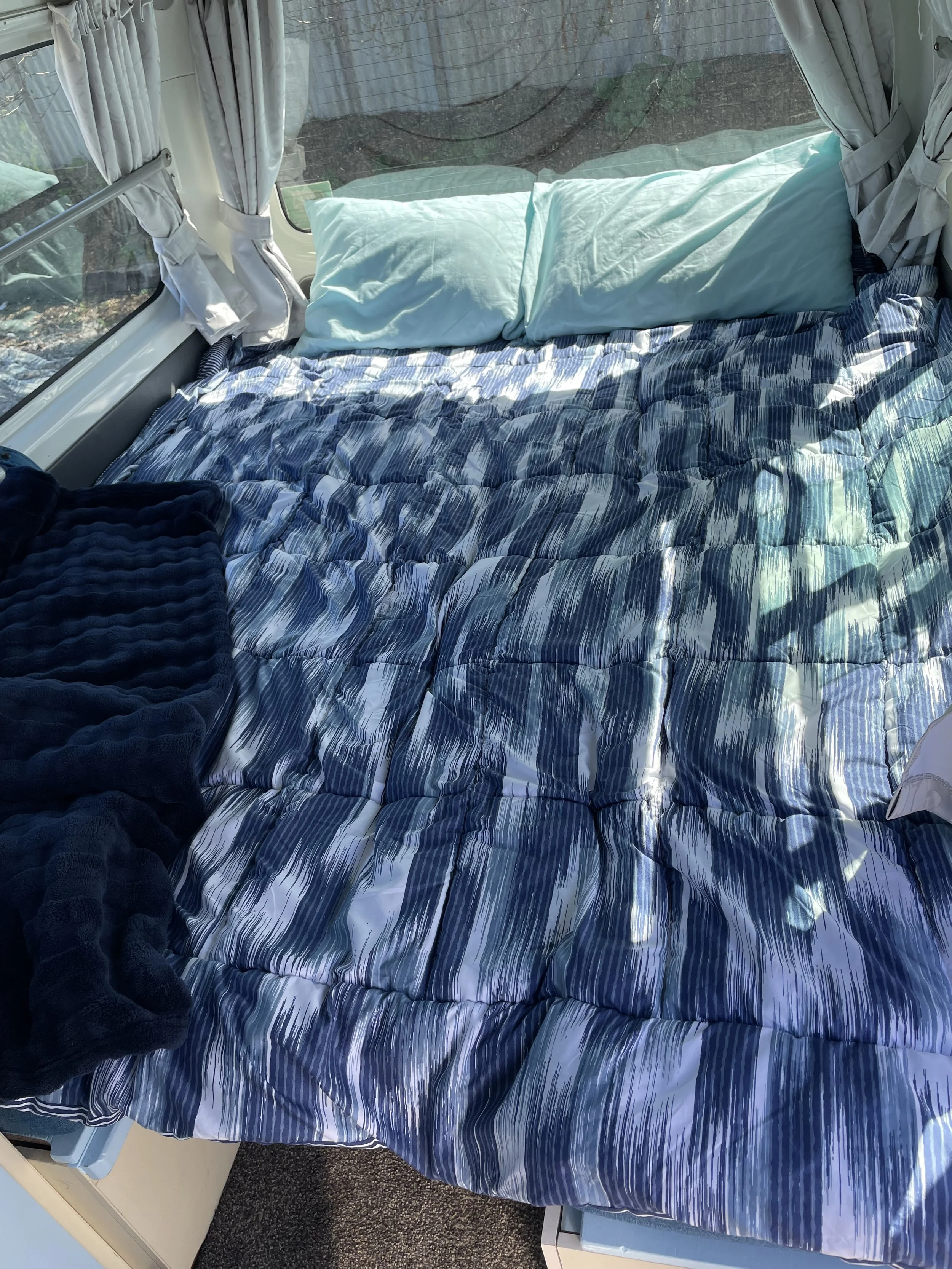 Bedroom inside a camper with a large window showing a scenic view of mountains, lake, and trees, with beige curtains and a blue bed covering.
