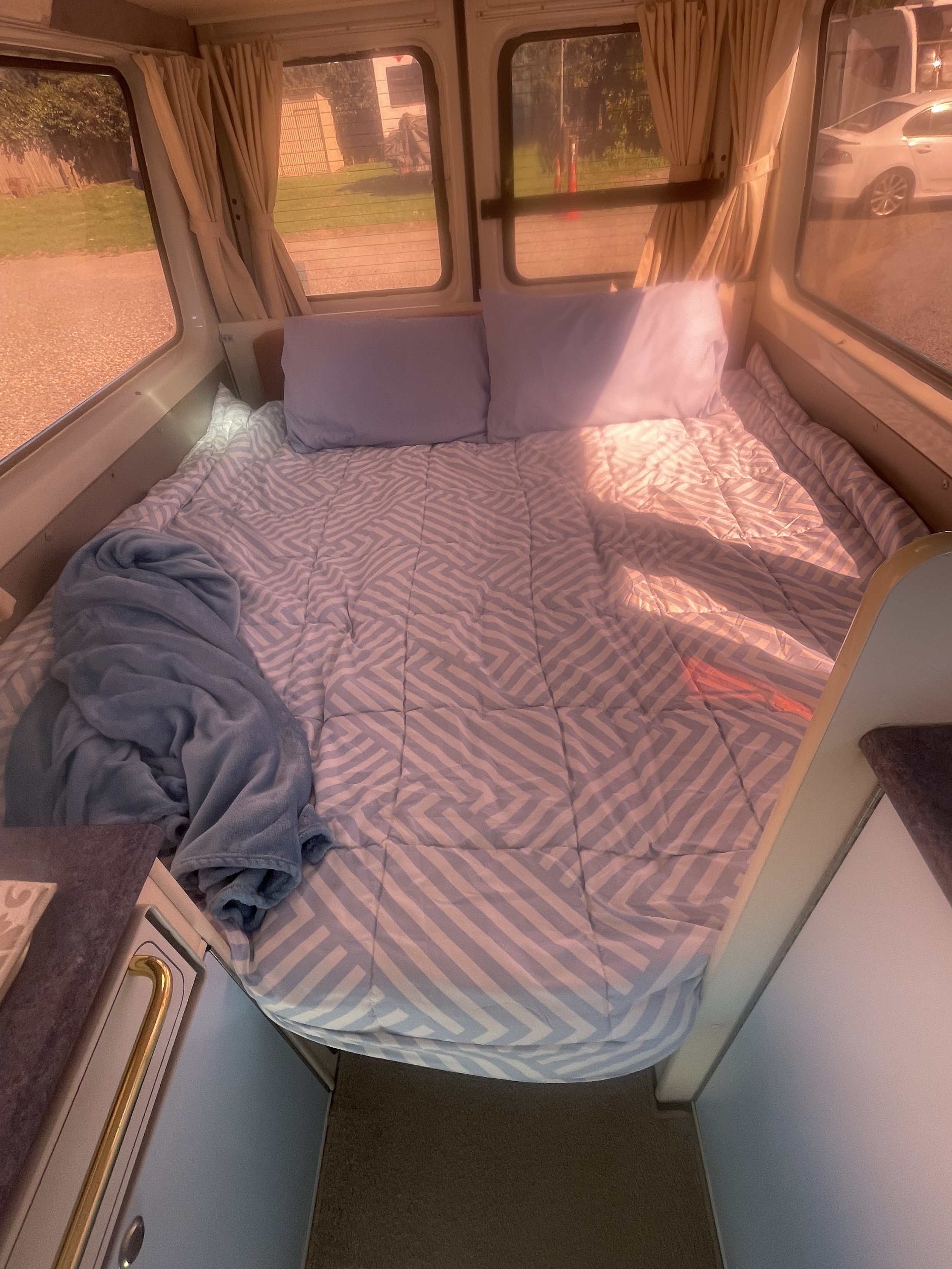 Bedroom inside a camper with a large window showing a scenic view of mountains, lake, and trees, with beige curtains and a blue bed covering.