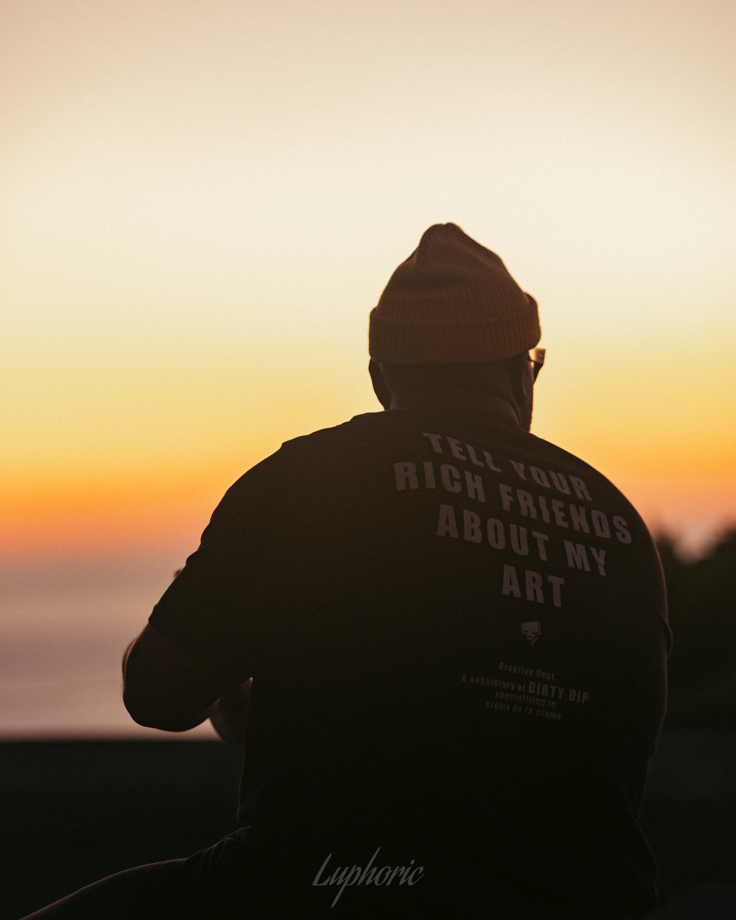 Silhouette of a person wearing a beanie and a black t-shirt with text, watching a sunset over the ocean with orange and pink sky.