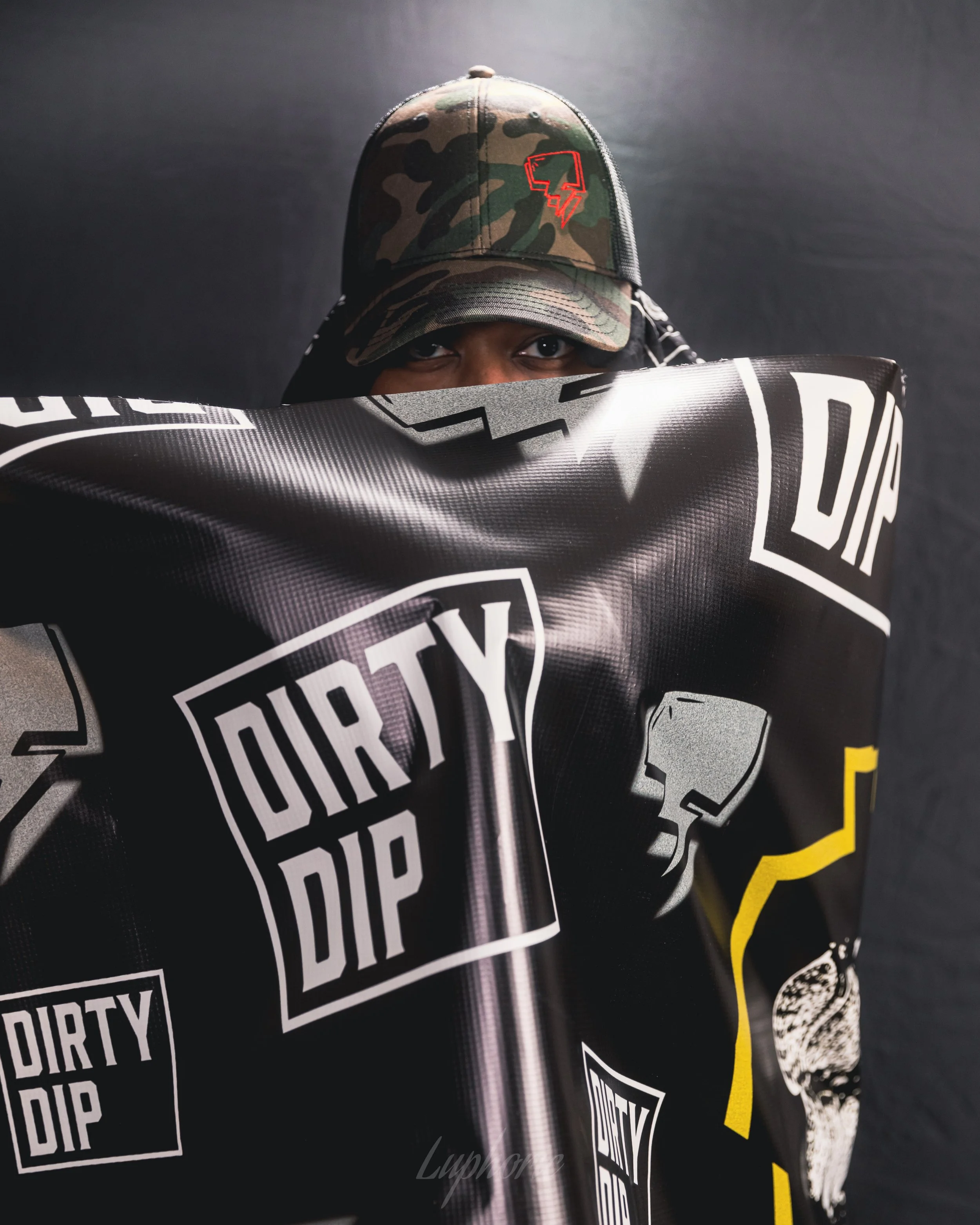 Person wearing a camouflage baseball cap with a red logo, partially hiding their face behind a black and white poster with logos and graphics.