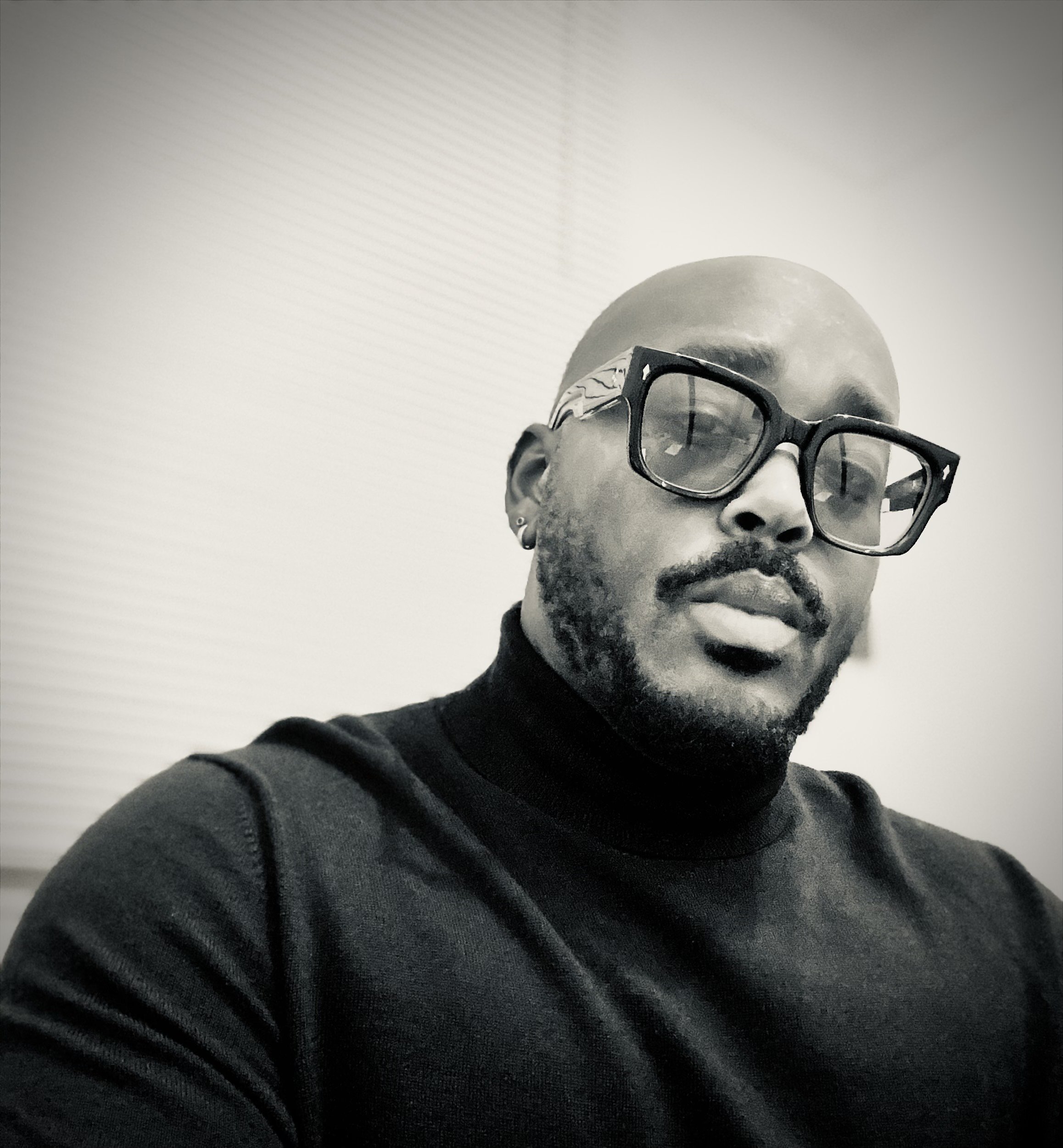 A man with a bald head, beard, and mustache wearing large black glasses and a black turtleneck sweater, sitting indoors with blinds in the background.