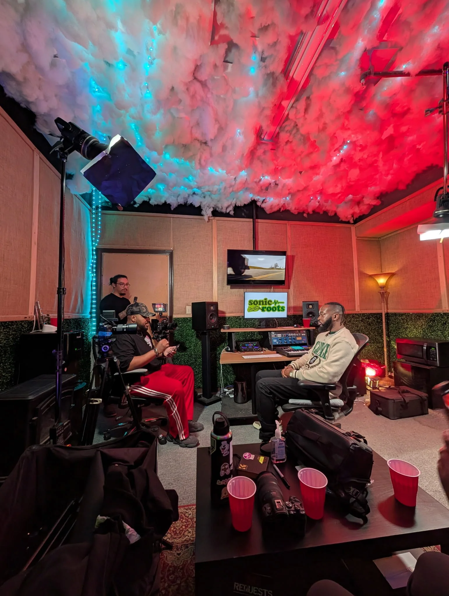 A recording studio with a ceiling decorated with cotton or fiber clouds illuminated by red and blue lights. Two men are seated facing each other, engaged in an interview, surrounded by cameras, microphones, and audio equipment. A monitor displays the logo 'Sonic Roots.' The room has beige soundproof walls, a floor lamp, and various tech gear and personal items on a table and the floor.
