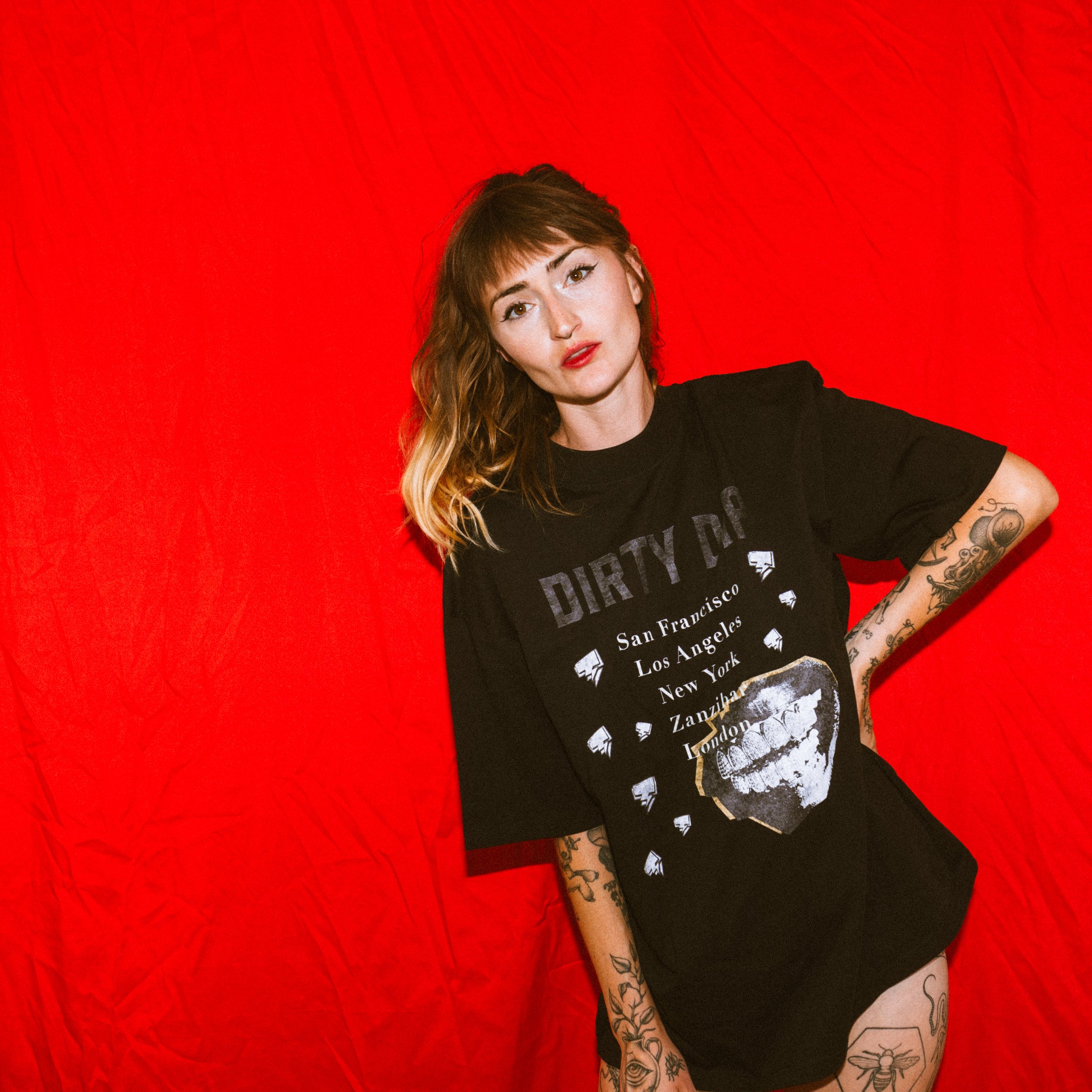 Young woman with shoulder-length hair and tattoos on her arms, wearing a black t-shirt with city names and graphic lips design, standing against a red backdrop.