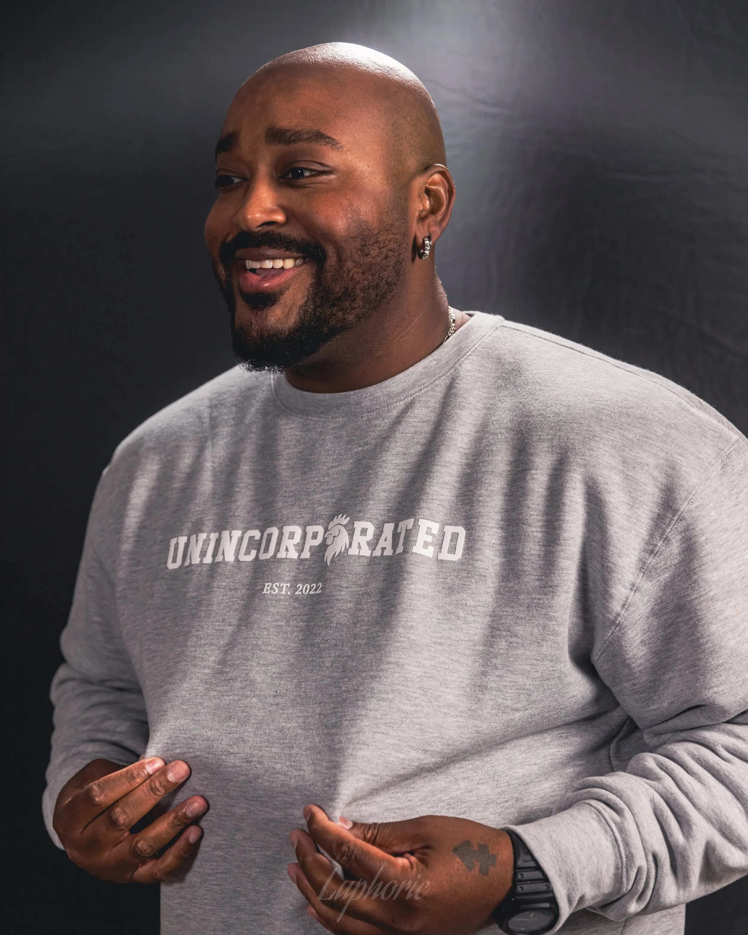 A smiling man with a shaved head and beard, wearing a gray sweatshirt that reads 'Unincorporated, Est. 2022,' standing against a black background.