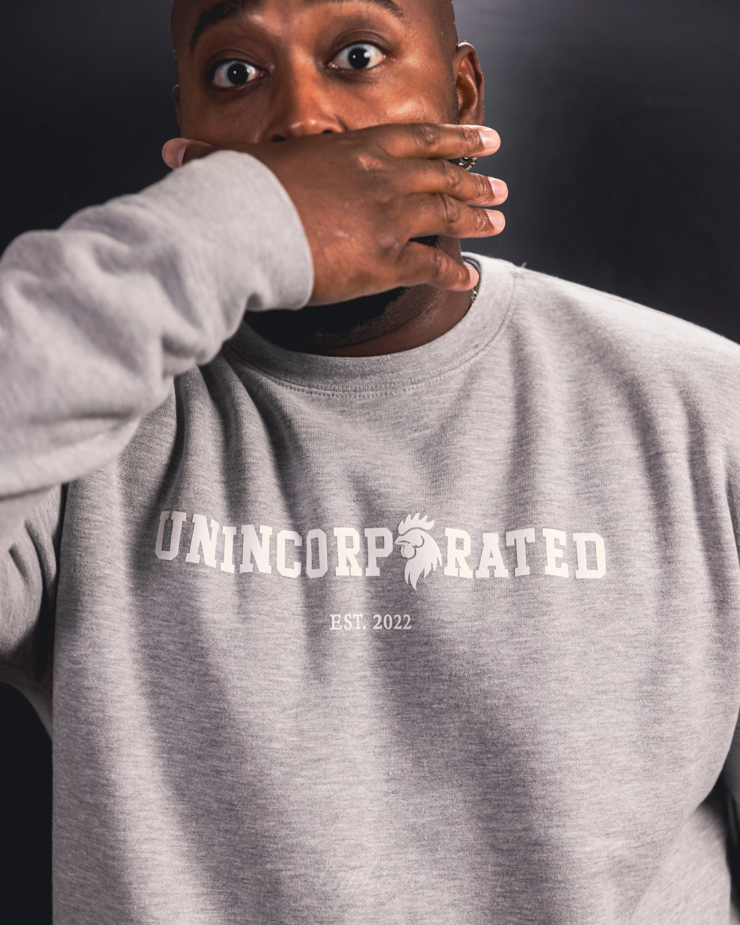 A person with dark skin holding their hand over their mouth, wearing a light gray sweatshirt with the word 'UNINCORPORATED' and a rooster logo, and the text 'EST. 2022' printed on it.