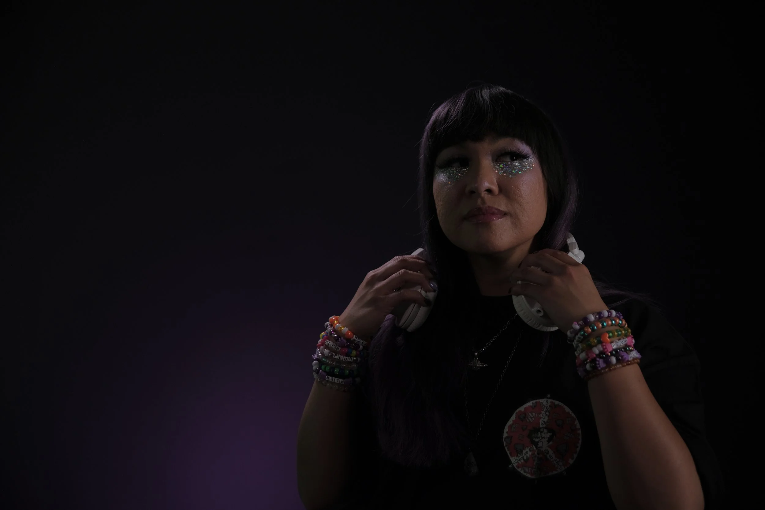 A woman wearing colorful bracelets and necklaces, with glitter on her face, holding headphones around her neck, in a dark setting with purple lighting.