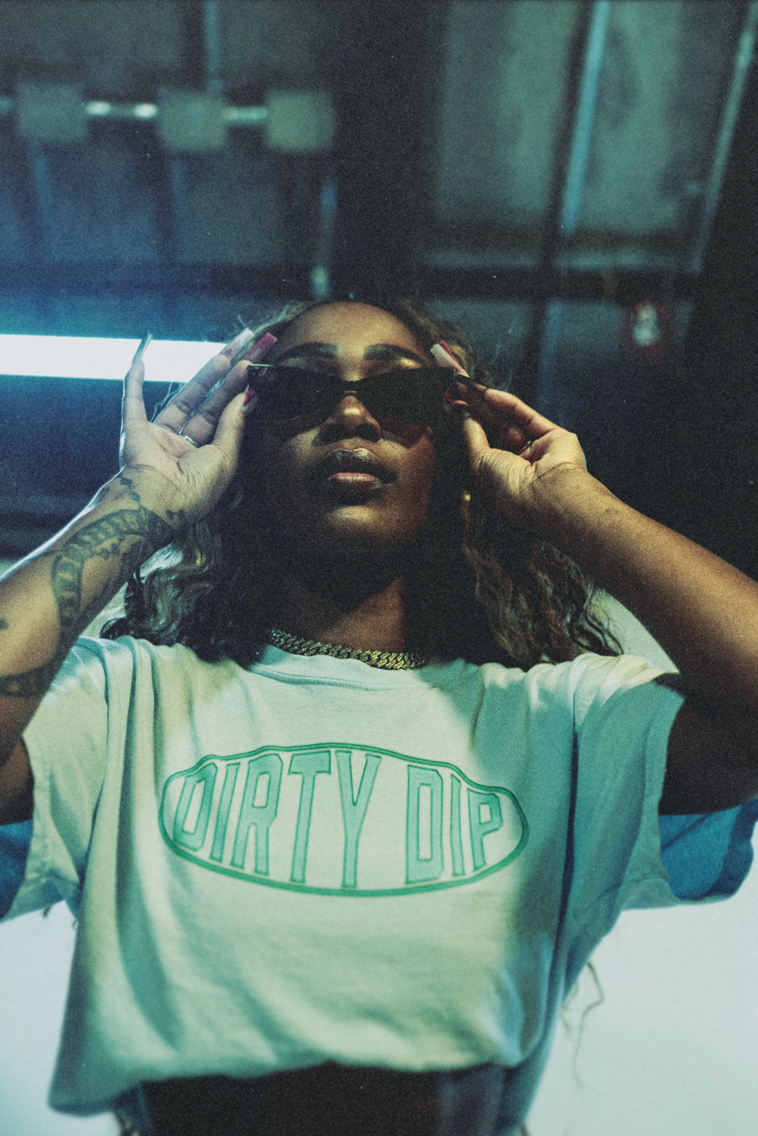 Woman wearing sunglasses, white t-shirt with 'DIRTY DIP' text, gold chain necklace, tattoos, and pink nail polish, posing indoors at night.