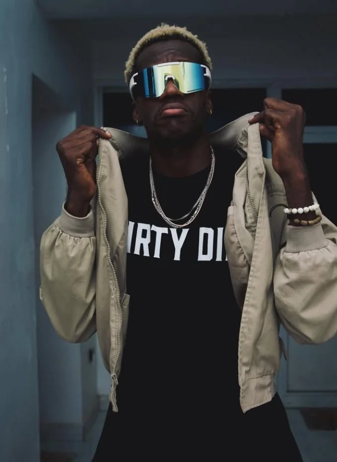 A man with blond hair wearing reflective sunglasses, a black T-shirt that says 'DIRTY DIG' in white, a beige jacket, and jewelry, posing with the jacket partially open.