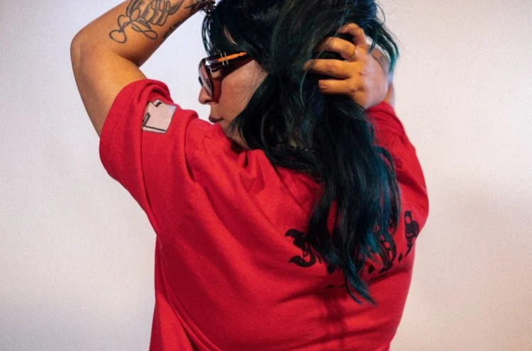 A woman with dark, blue-tinted hair wearing sunglasses and a red shirt, standing against a plain wall, with one arm raised and hand in her hair, turned to the side.