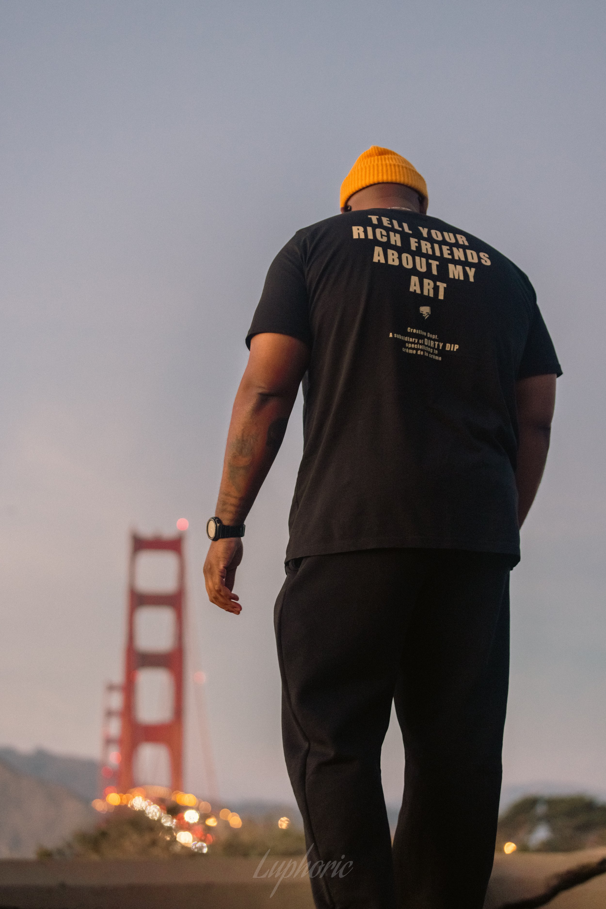 A person wearing a black T-shirt with the message "Tell your rich friends about my art" and a bright yellow beanie, standing in front of the Golden Gate Bridge during dusk.