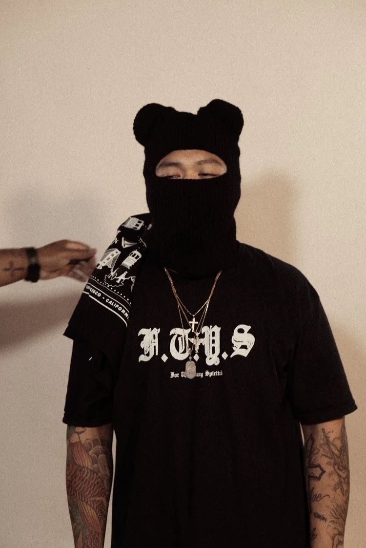 A person wearing a black ski mask with bear ears, black T-shirt with white Gothic lettering, gold chains, and tattoos on their arms, standing against a plain wall while someone else adjusts their shoulder.
