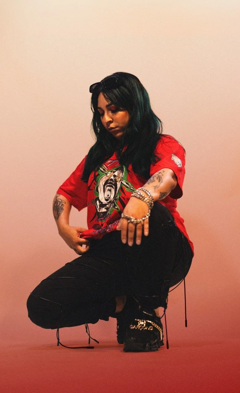 A woman with black hair, tattoos, and wearing sunglasses on her head squats against a plain pink background. She is dressed in a red graphic T-shirt, black pants, and black shoes with embellishments.