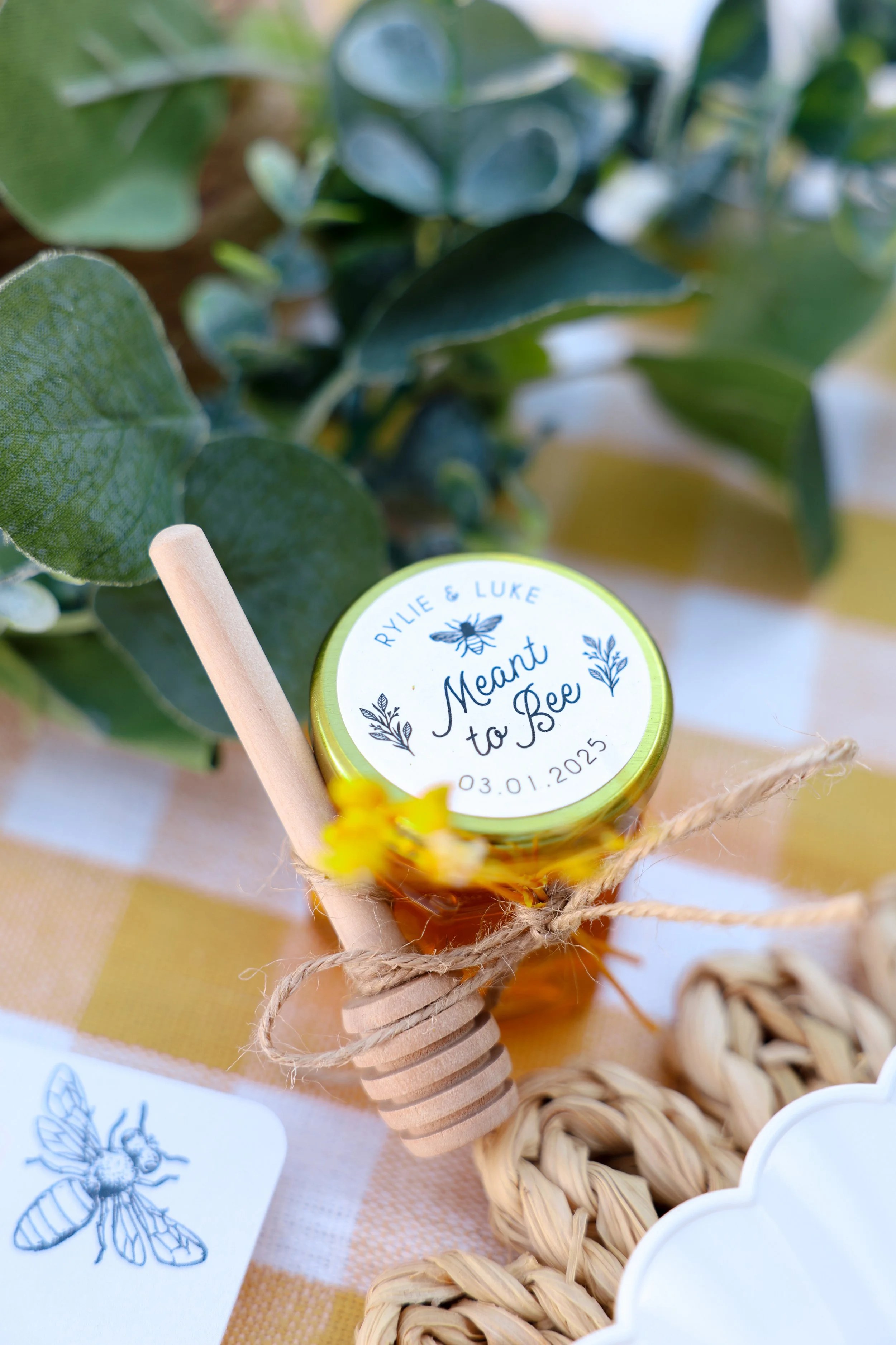 A small jar of honey with a personalized label that reads 'Rylie & Luke, Meant to Bee, 03.01.2025' along with a bee illustration, placed on a woven mat with greenery around it.
