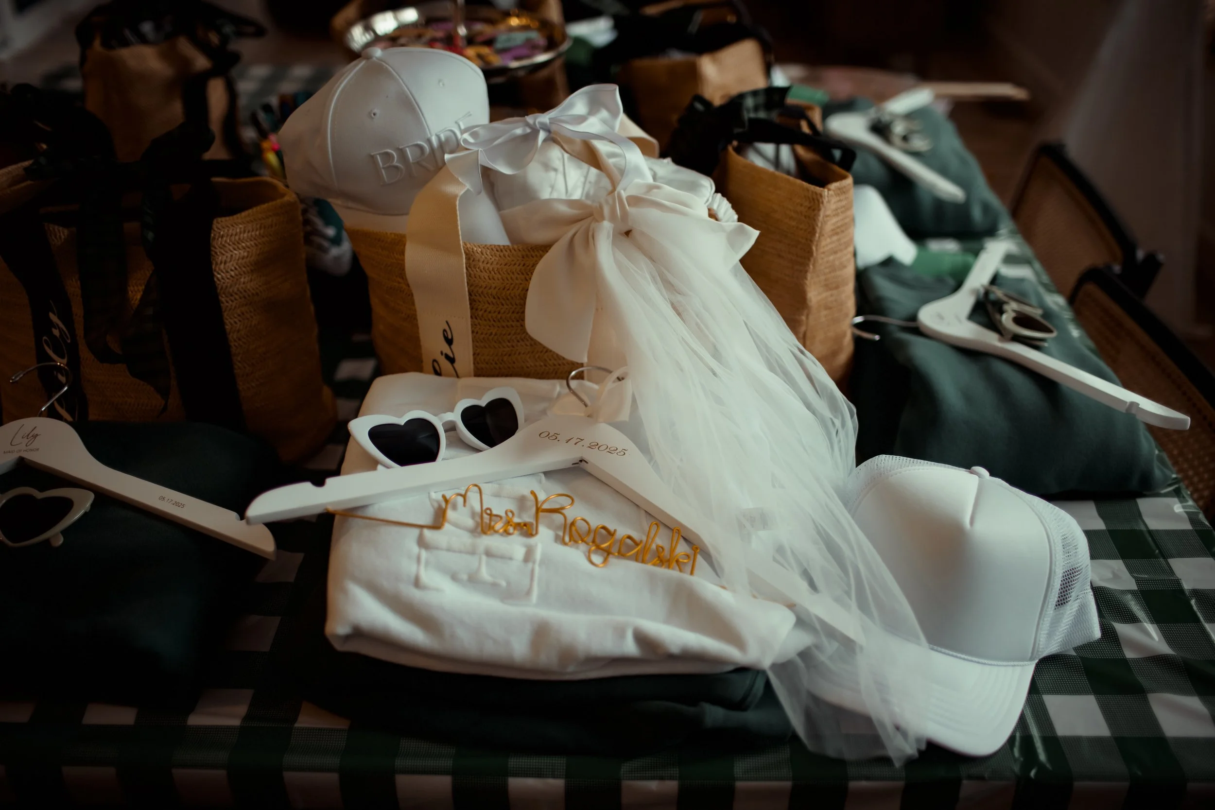 Wedding accessories including white sunglasses, a white cap with 'BRIDE' embroidered, a white veil, a white cap, a white t-shirt with 'Mrs. Rogelki' written in gold wire, and a white hanging tag with a date on it, on a checkered tablecloth.