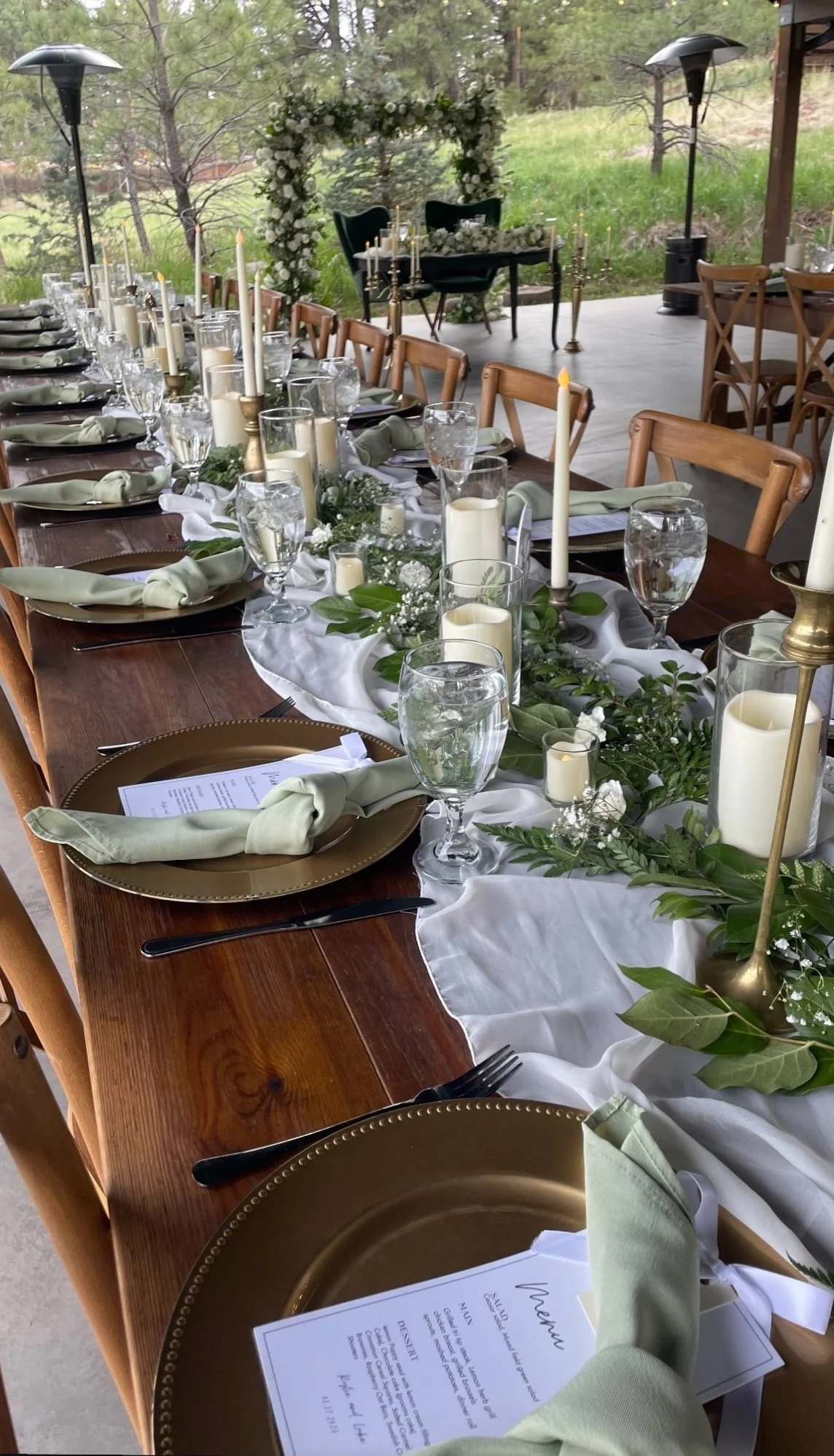 A long wooden table decorated for a wedding reception, with greenery, white flowers, candles, and gold accents, set outdoors on a covered patio surrounded by trees.