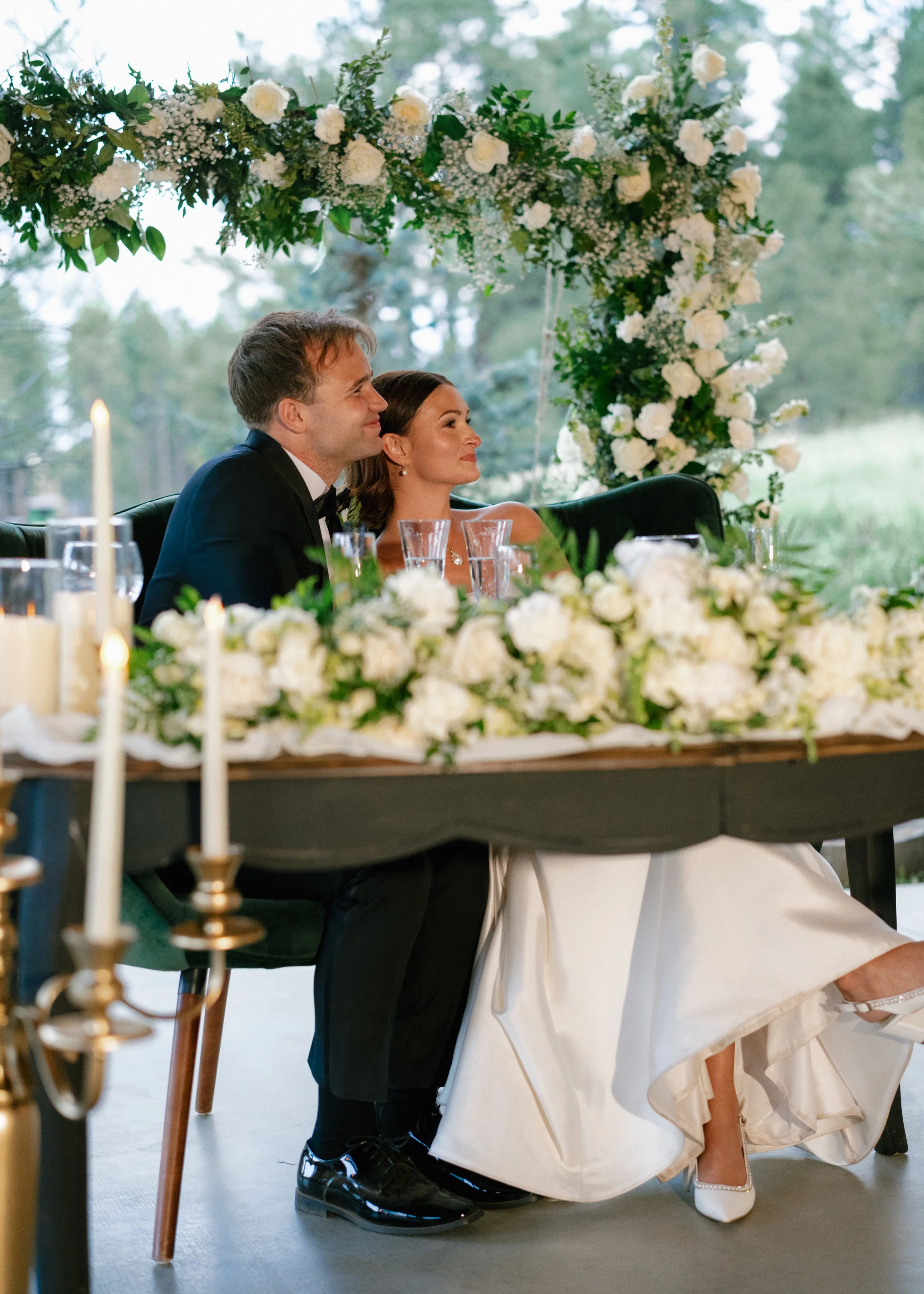 Two people sitting at a wedding reception table, a man and woman, surrounded by white floral arrangements and candles, with a natural outdoor background.