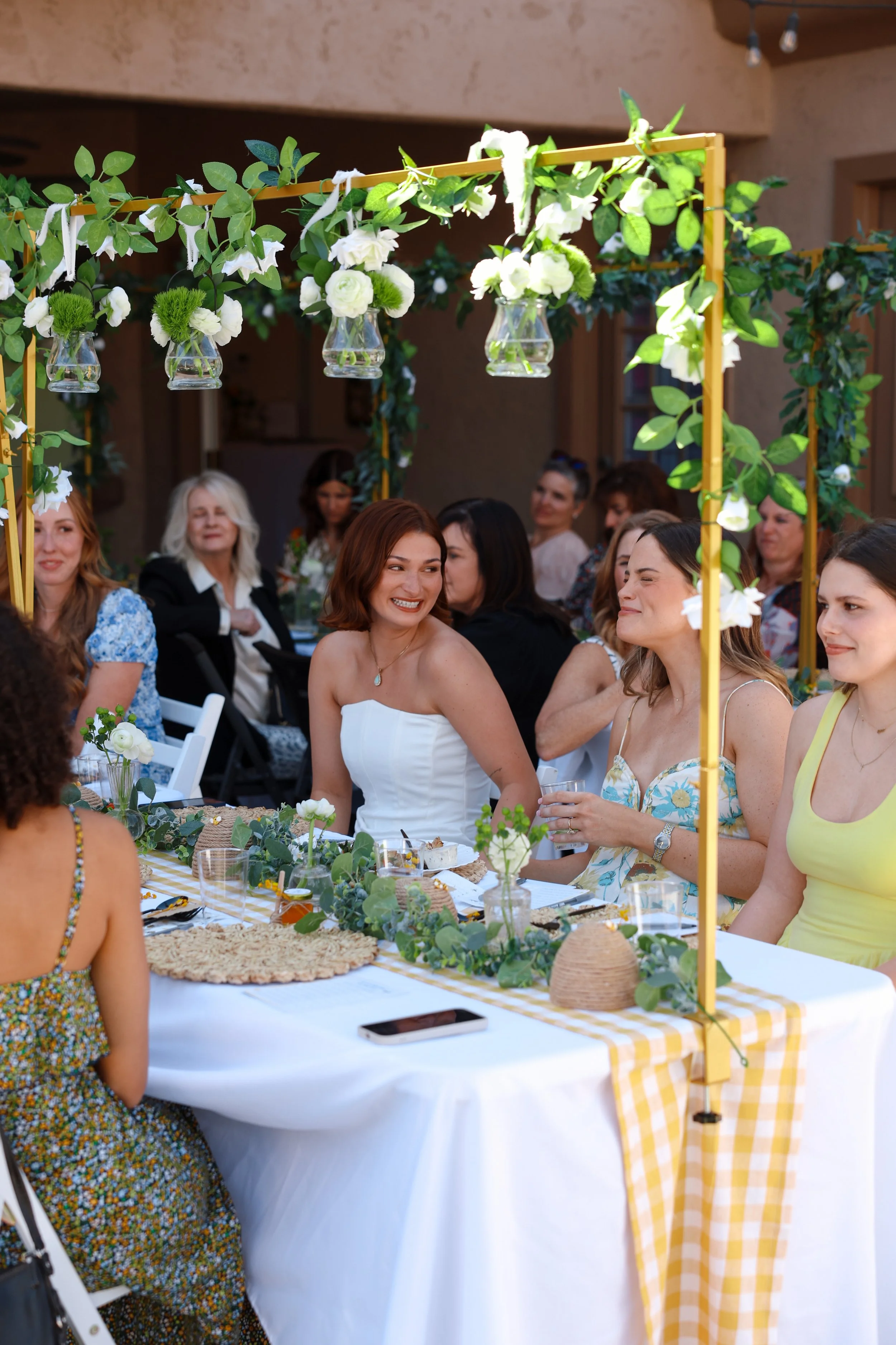 Group of women enjoying a celebration at a decorated outdoor table with greenery and hanging vases with white flowers.