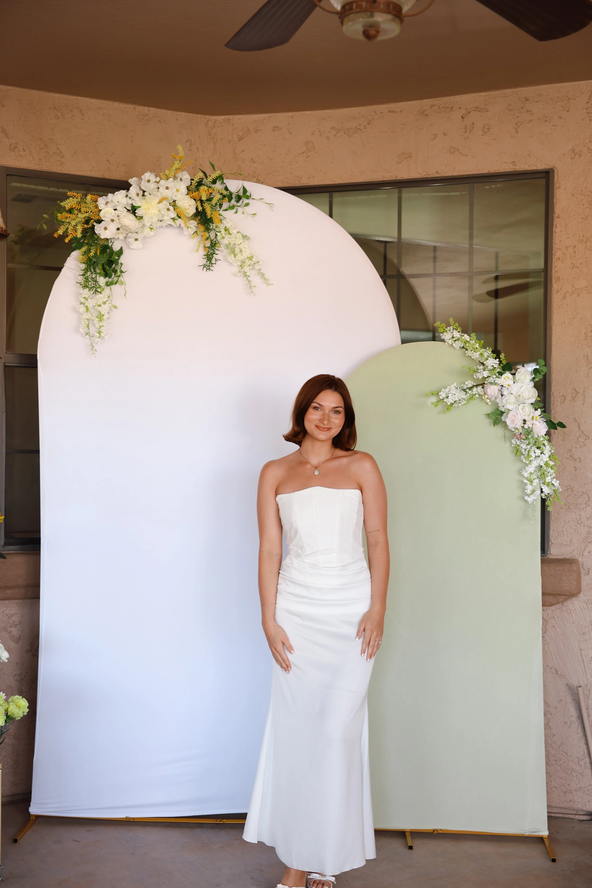 A young woman in a strapless white dress standing in front of decorative white and light green backdrops decorated with white and yellow flowers.