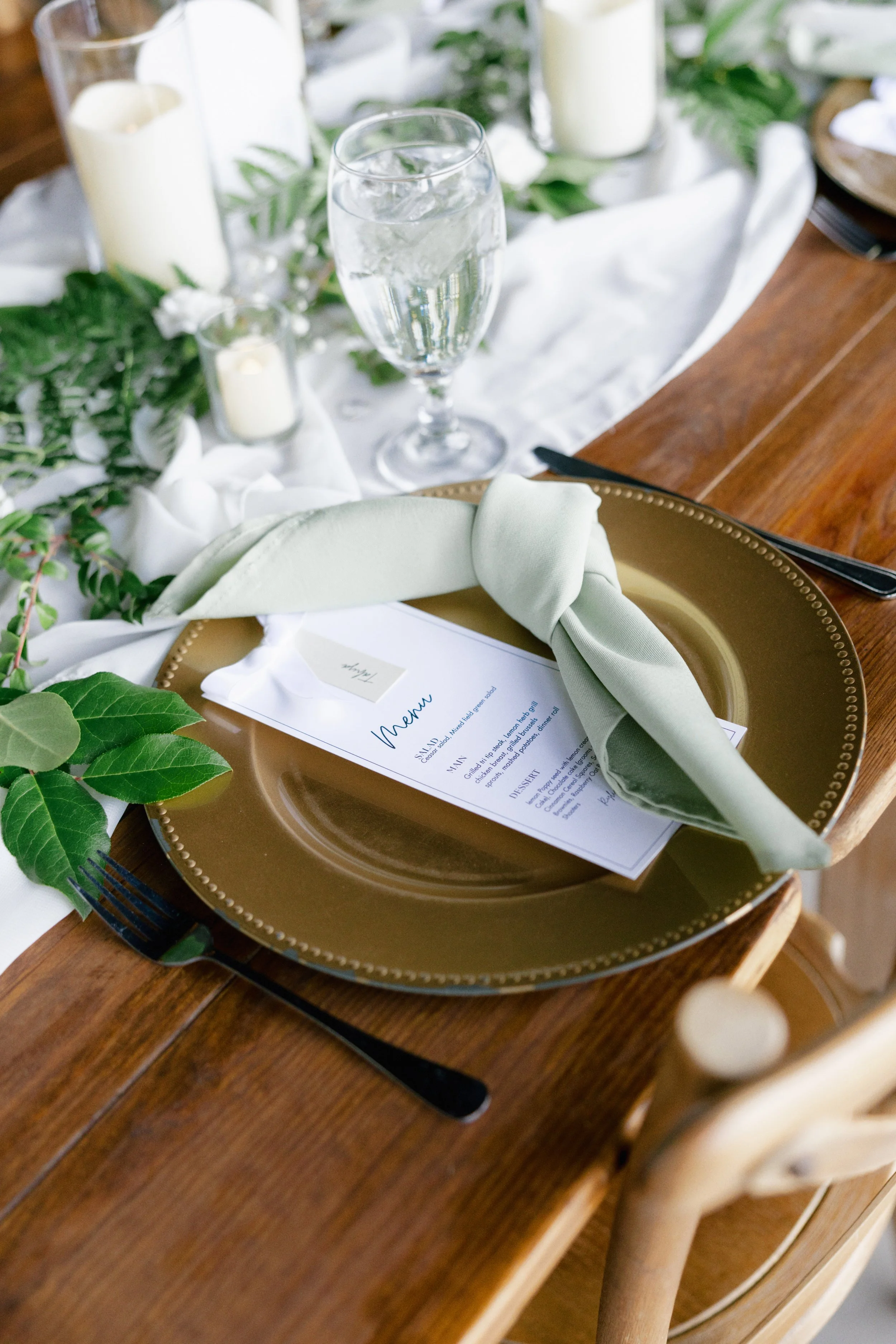 Elegant table setting with a gold charger plate, a green cloth napkin, a menu card, and floral and candle decor in the background.