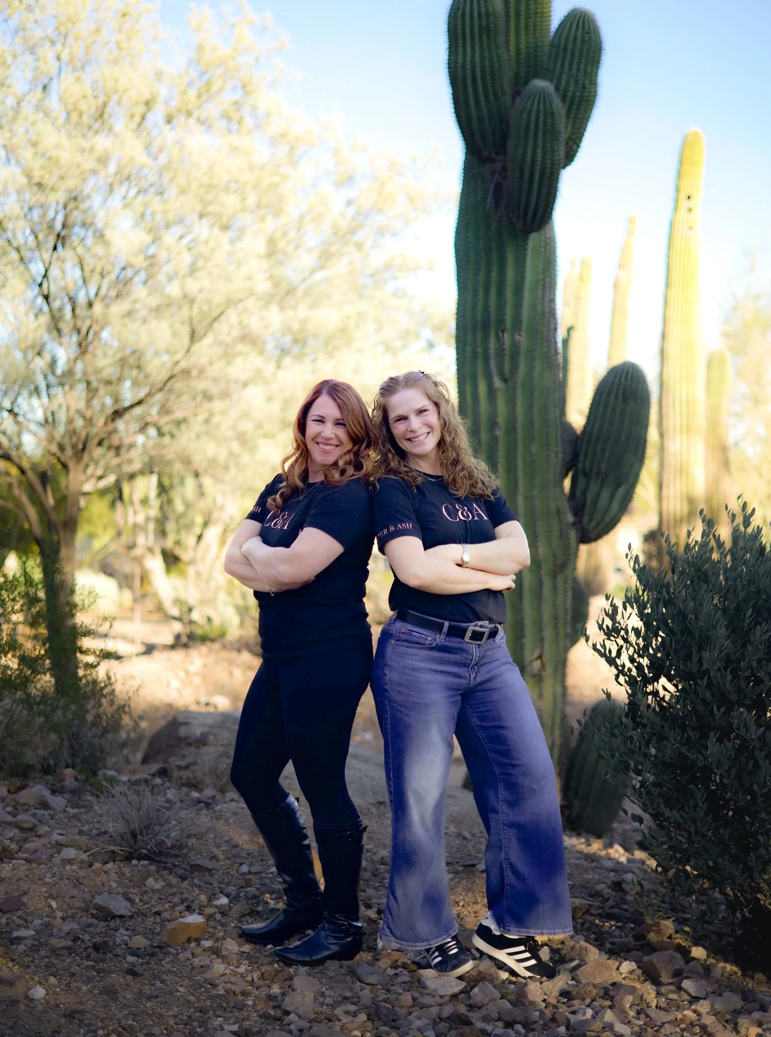 Two women standing outdoors in a desert landscape with large cacti and trees, smiling and facing the camera with arms crossed, wearing matching black T-shirts and jeans.