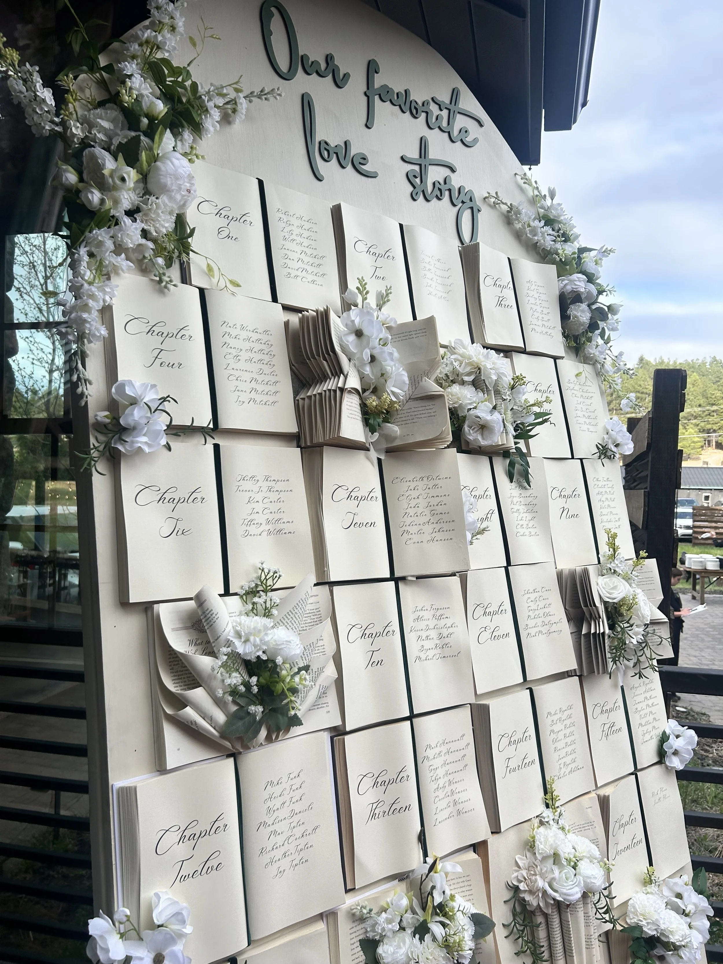 Display of handwritten guest book entries arranged by chapter numbers, decorated with white flowers and greenery, with the title 'Our favorite love story' at the top.