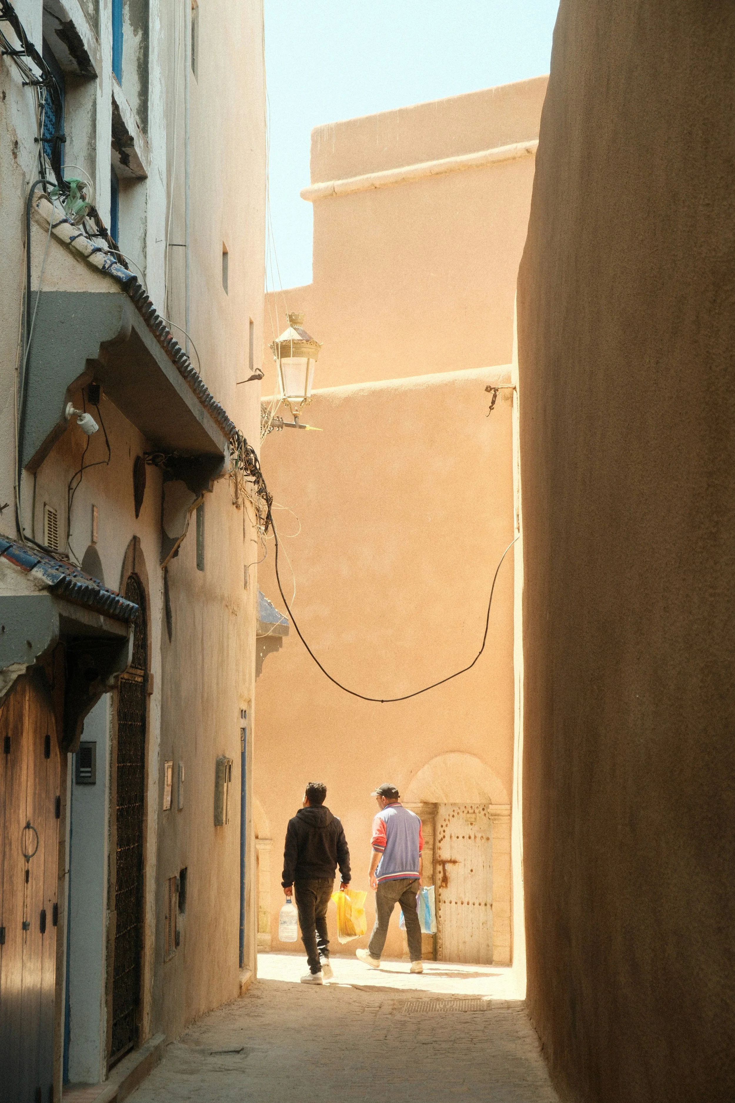 Al Seef Dubai Wayfinding. Two people walking down a narrow alleyway carrying plastic bags in a desert-like town with beige and tan buildings and a clear blue sky.