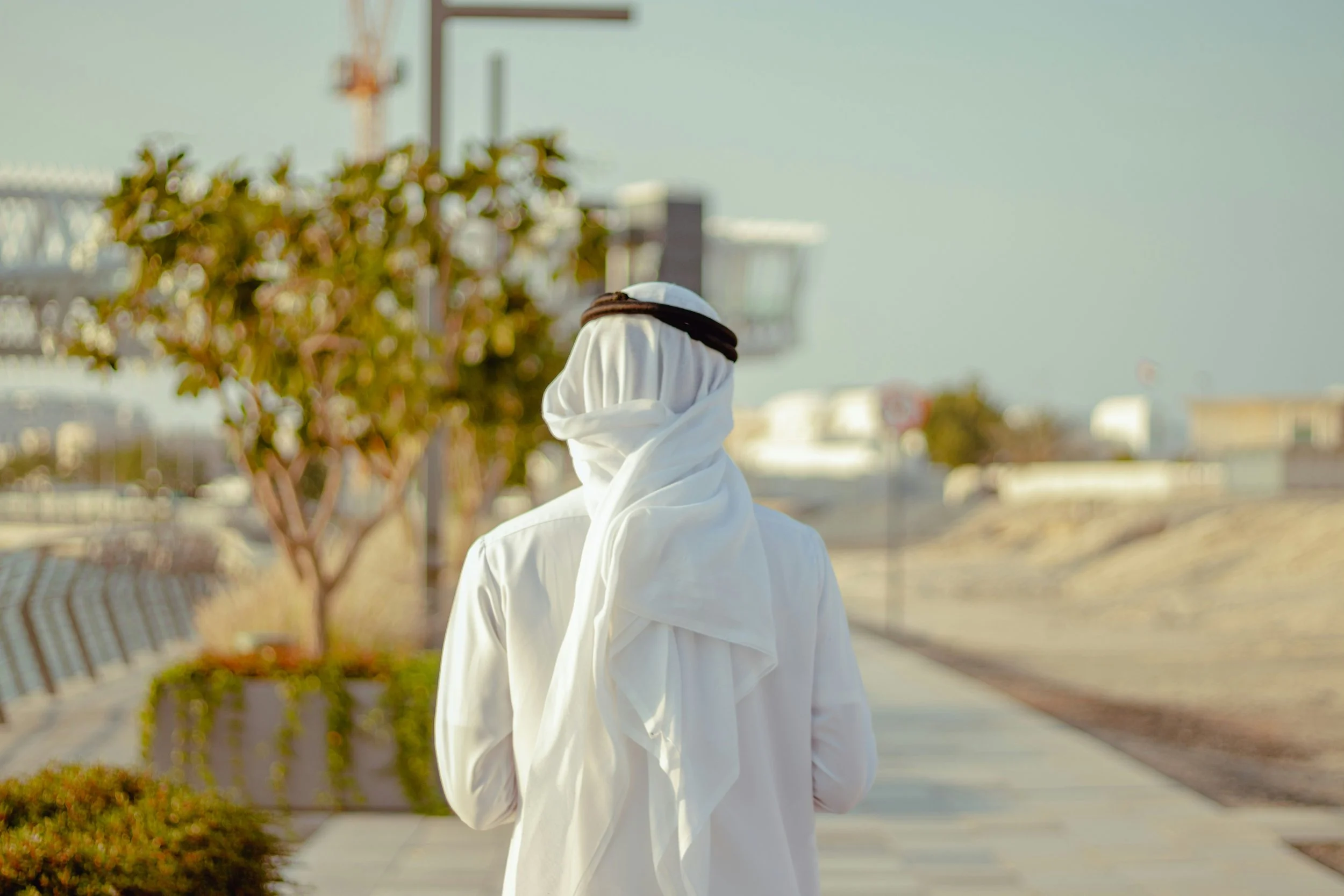 Visitor Experience Design. Emirati man wearing traditional Middle Eastern attire, including a white thobe and a black and white ghutra, walking outdoors in Dubai on a paved path with trees and a cloudy sky in the background.