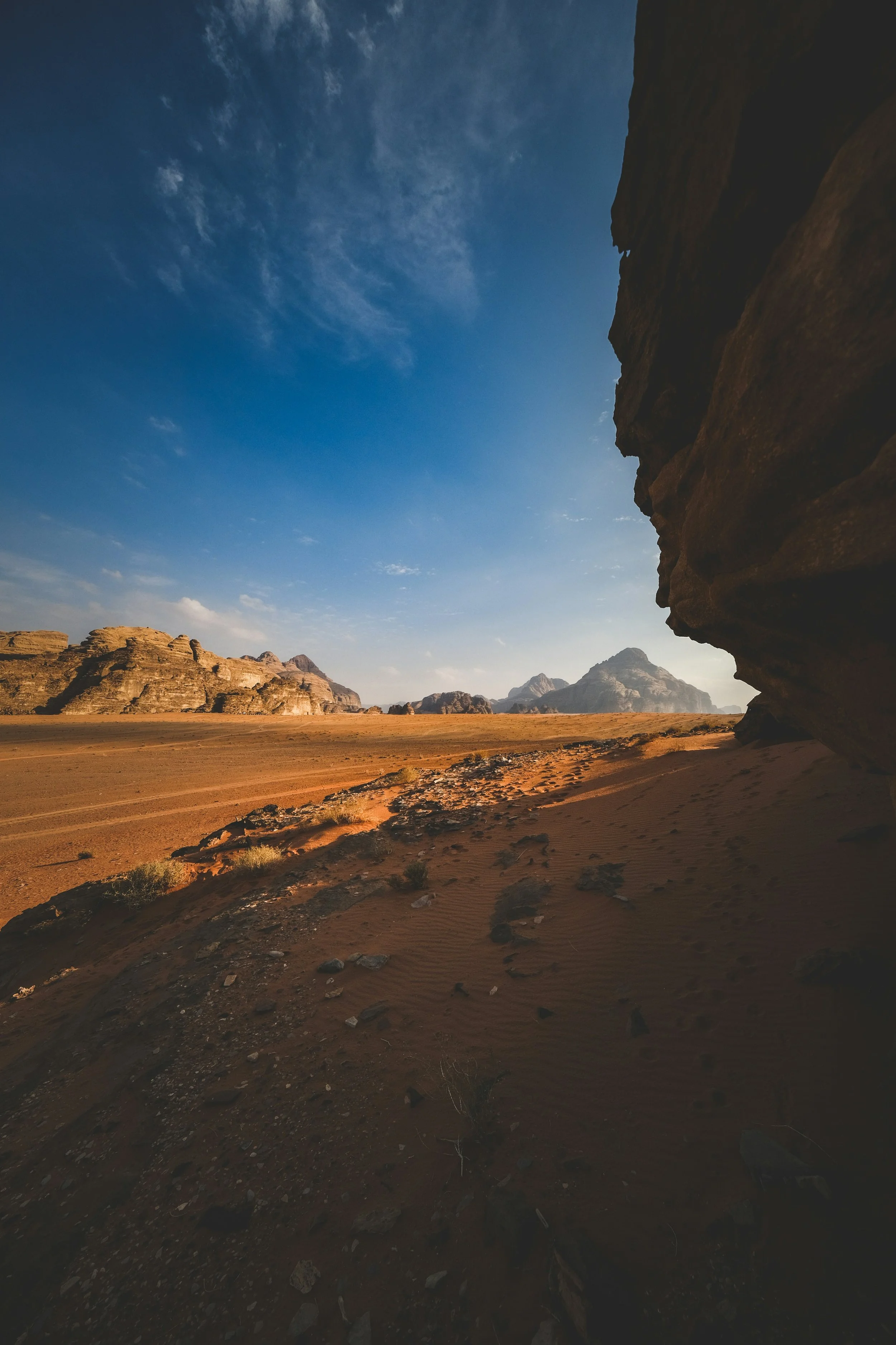 A view of a desert landscape with sandy terrain, rock formations, and mountains in the distance under a blue sky with scattered clouds.