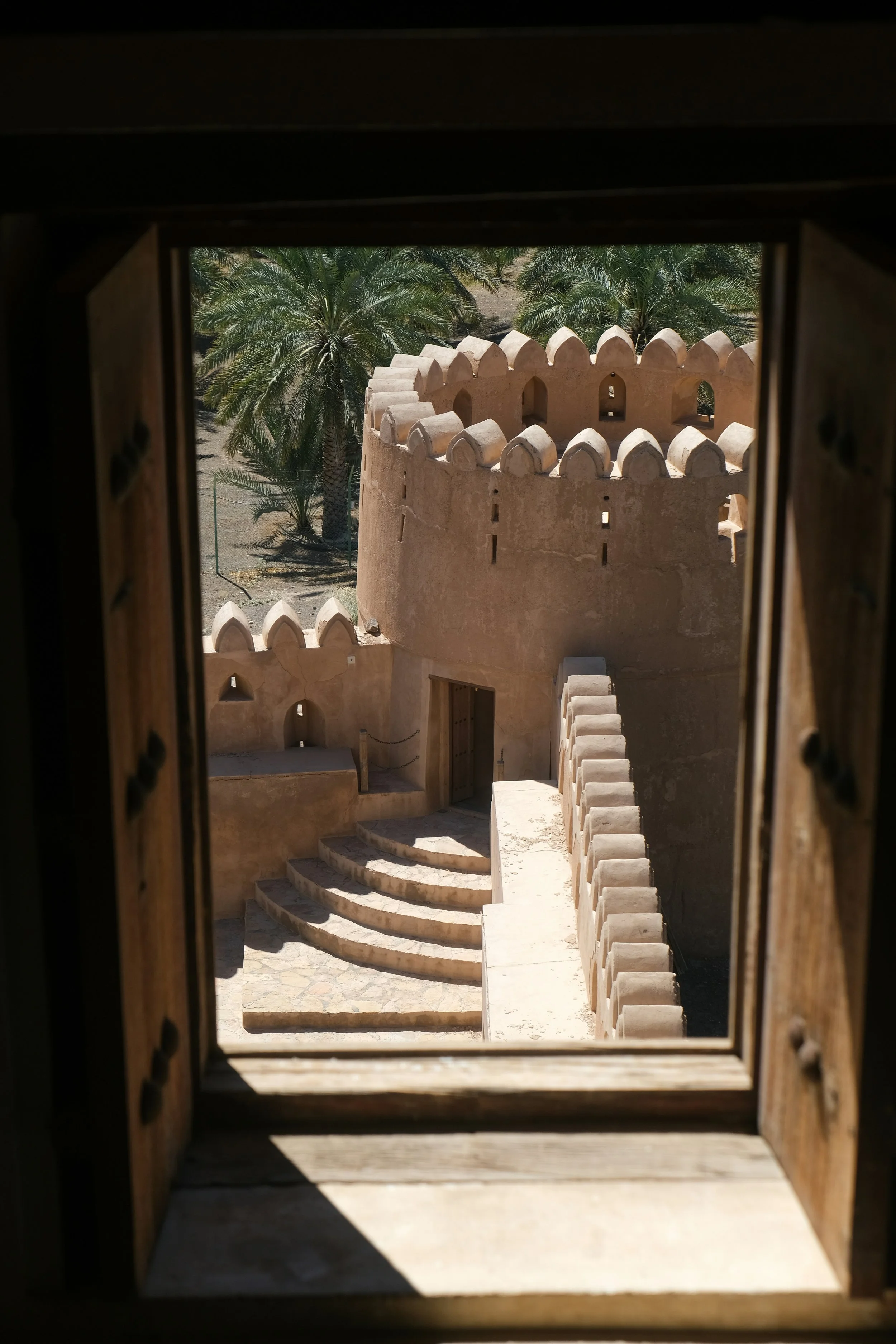Wayfinding. View of a traditional Oman desert fortress with rounded towers and battlements, framed by a window with wooden shutters, amid palm trees.