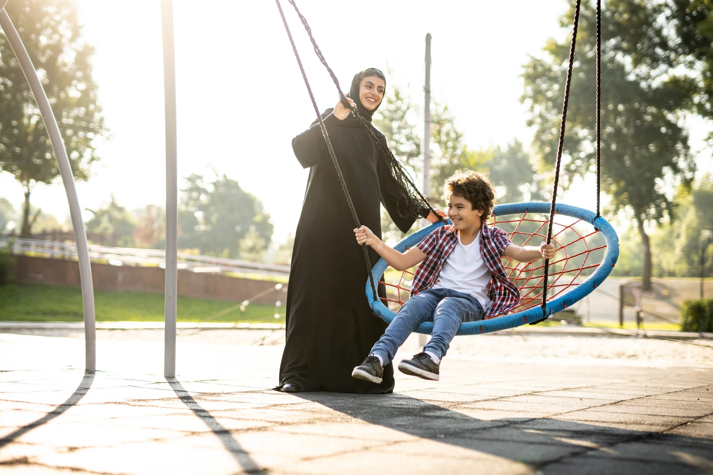 Community Placemaking. Arabic mother in a black shawl and a young boy with curly hair sitting on a blue swing at a Dubai park during sunny weather.