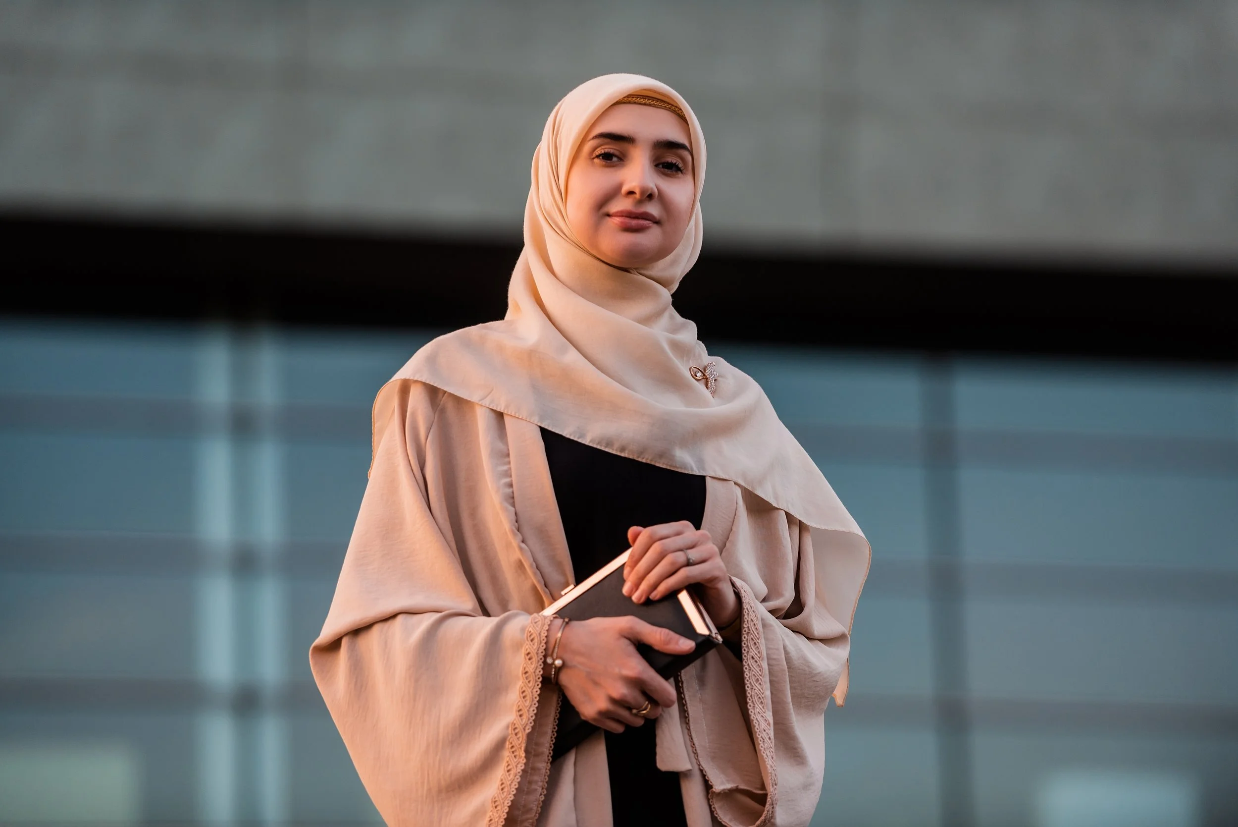 Visitor Experience Strategy. Arabic woman wearing a beige hijab and beige modest clothing, holding a black notebook, standing outdoors in front of a modern building with glass panels.