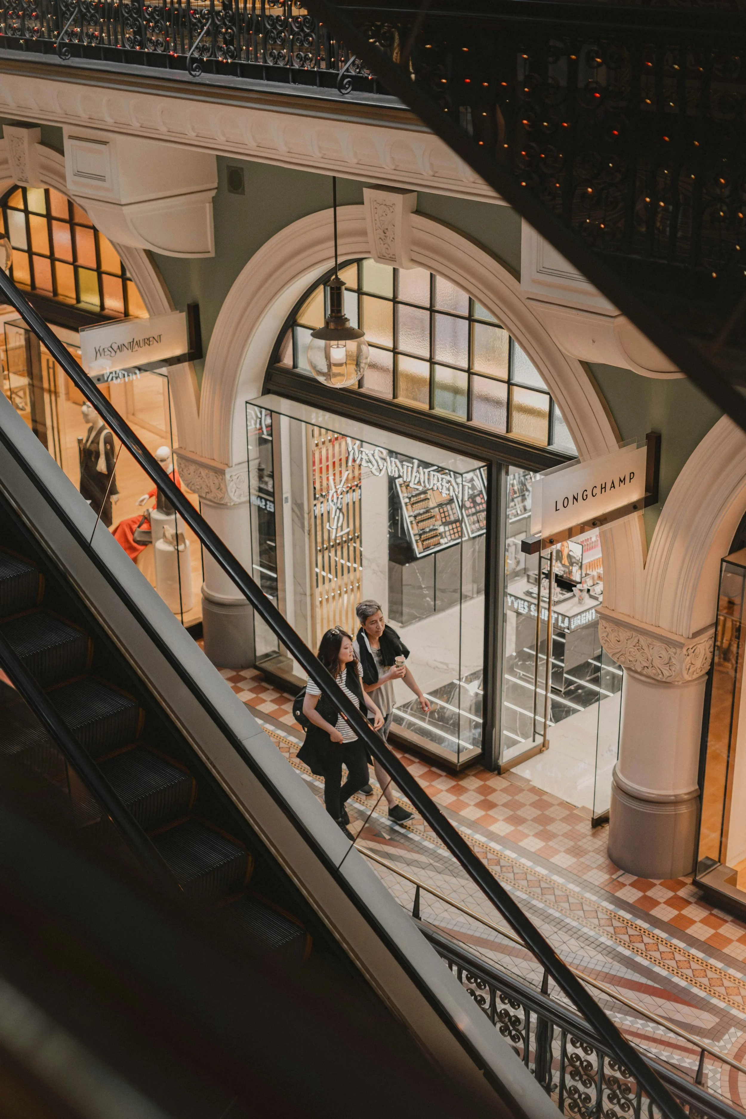 Retail Wayfinding. Two women walking down an escalator inside a shopping mall with luxury designer stores, including Yves Saint Laurent and Longchamp, visible through glass storefronts.