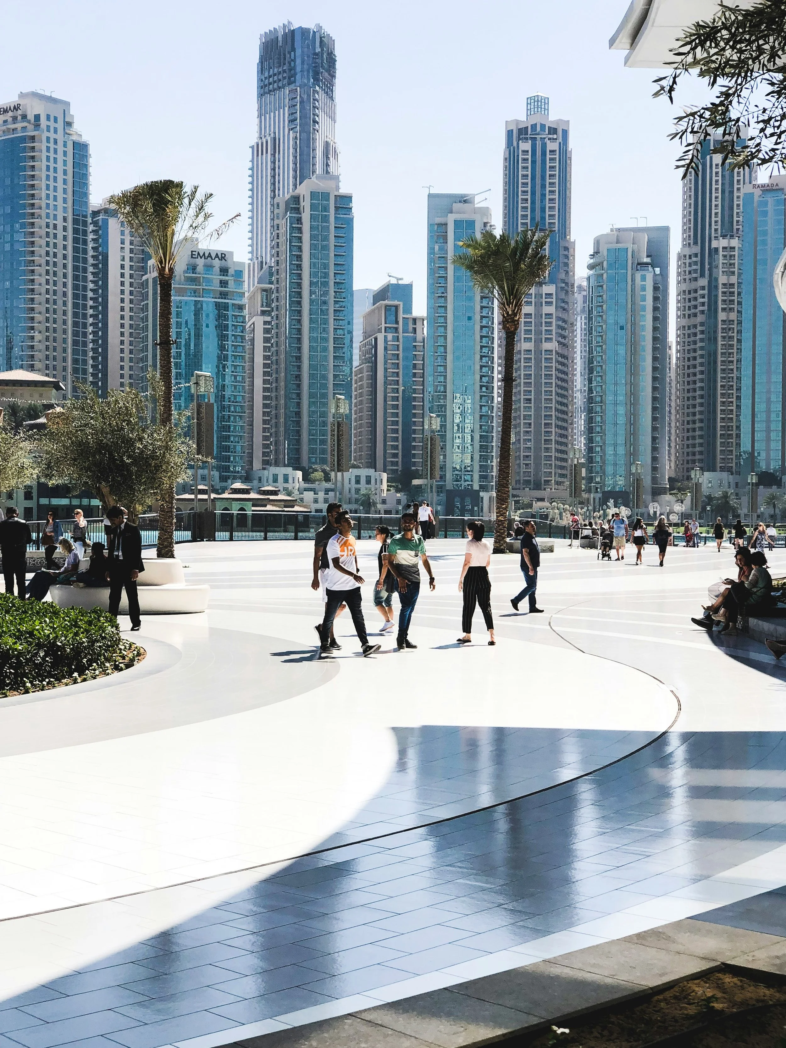 Public Realm Wayfinding. Downtown Dubai cityscape with tall skyscrapers, palm trees, and people strolling on a sunny outdoor plaza.