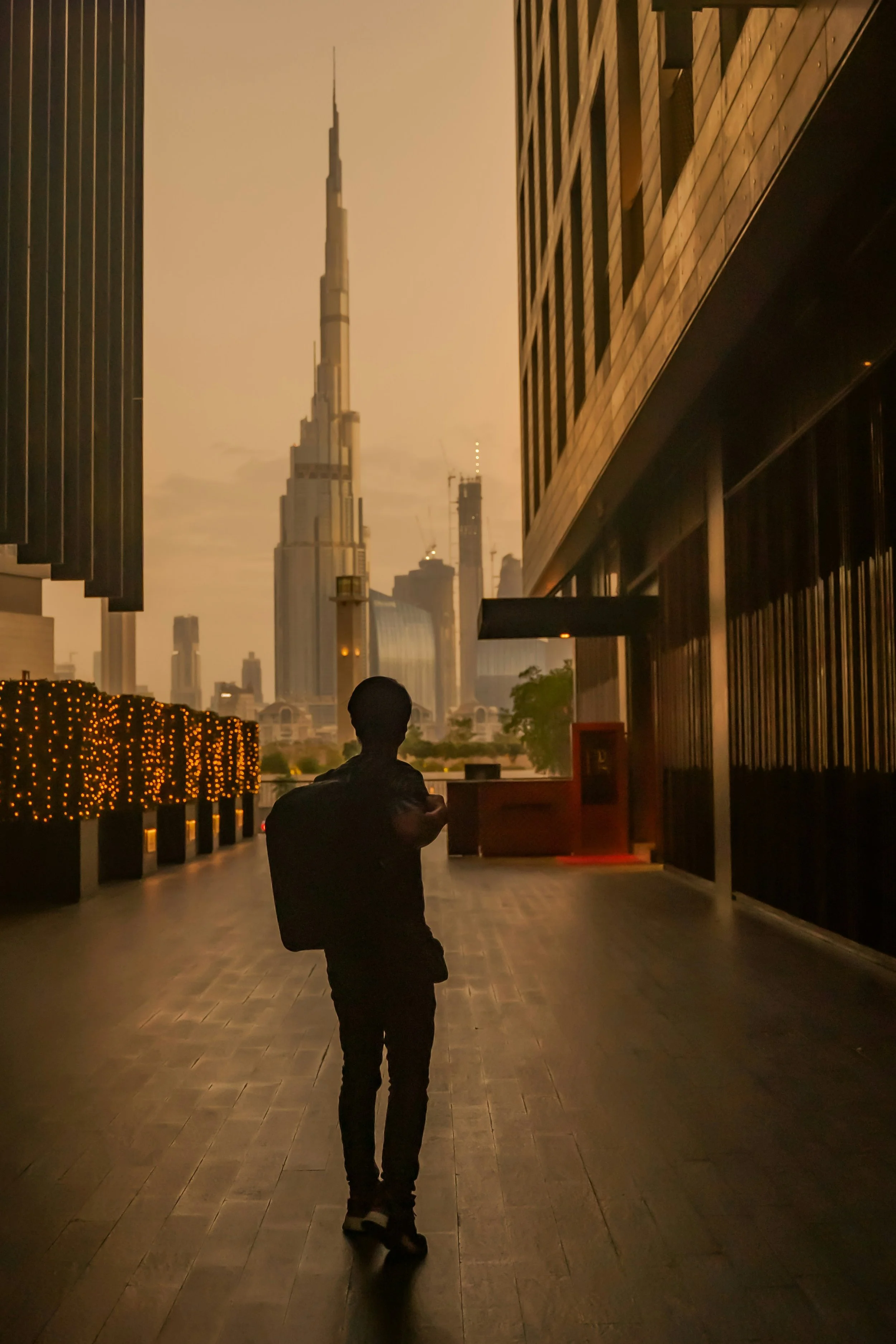 A silhouette of a person standing on a tiled pedestrian pathway at night, with city skyscrapers, including the Burj Khalifa, in the background under an orange-tinted sky.