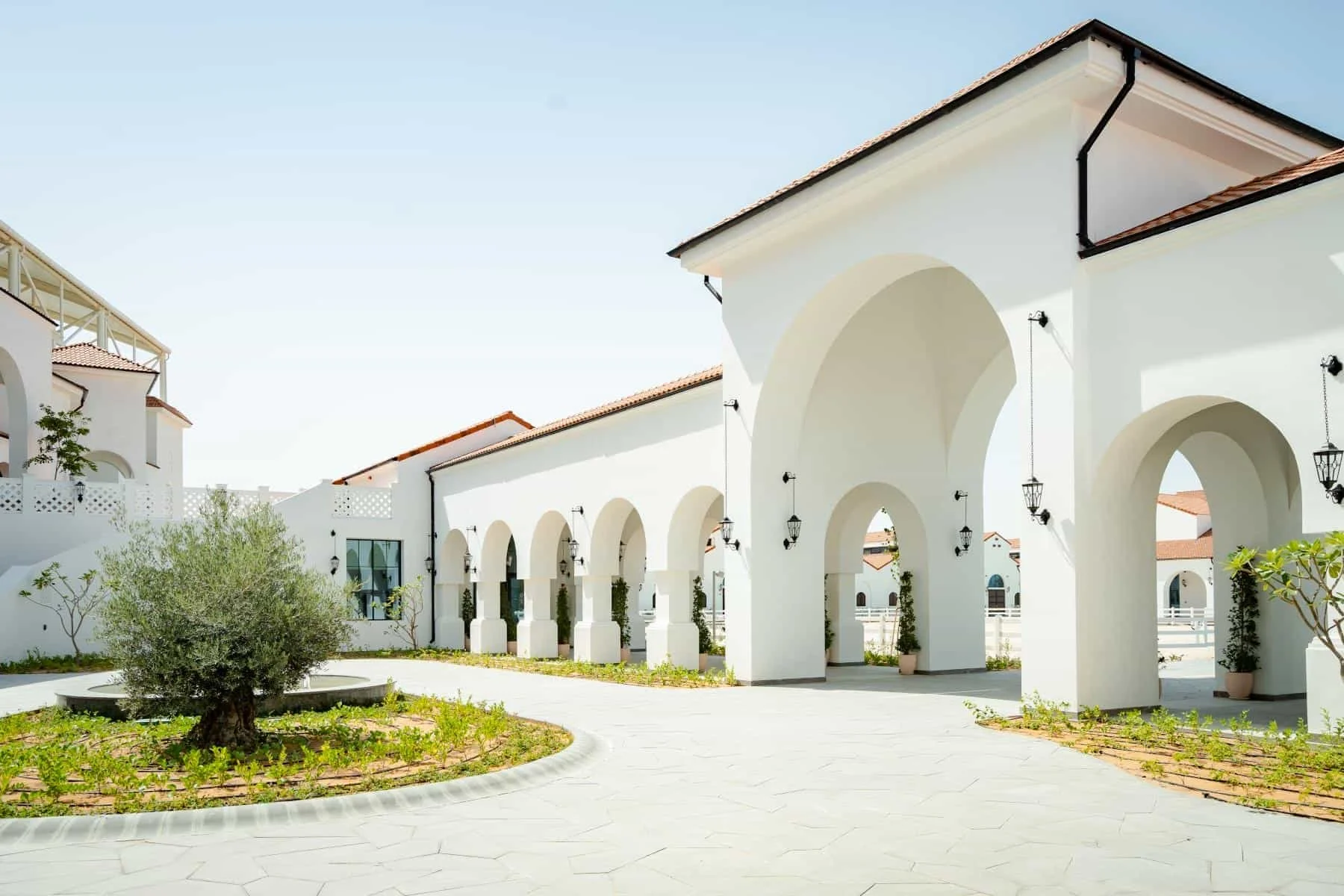 A white building with arched walkways, black lantern-style light fixtures, and a red-tiled roof, with a small garden and plants in the foreground under a clear blue sky.