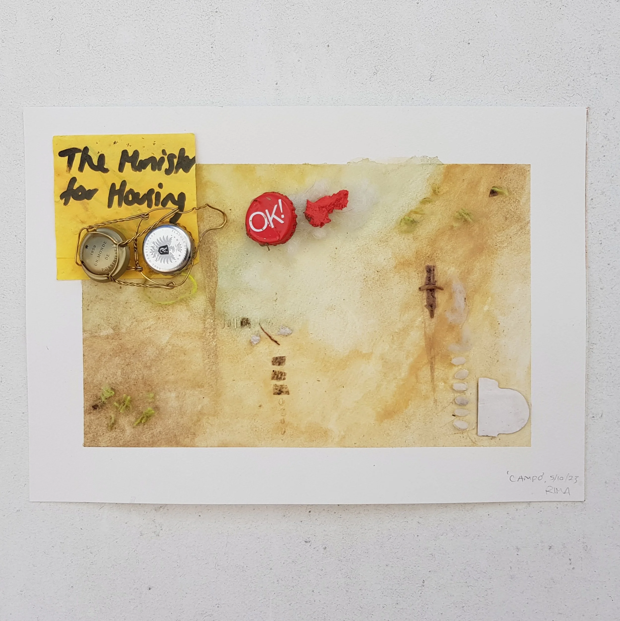 Mixed media artwork titled 'Camp 5/10/23 RIMA' featuring a watercolor background with small objects such as bottle caps, a red wrapper, small rocks, and a yellow note with handwritten text, arranged to resemble a desert scene with a cross and a cup.