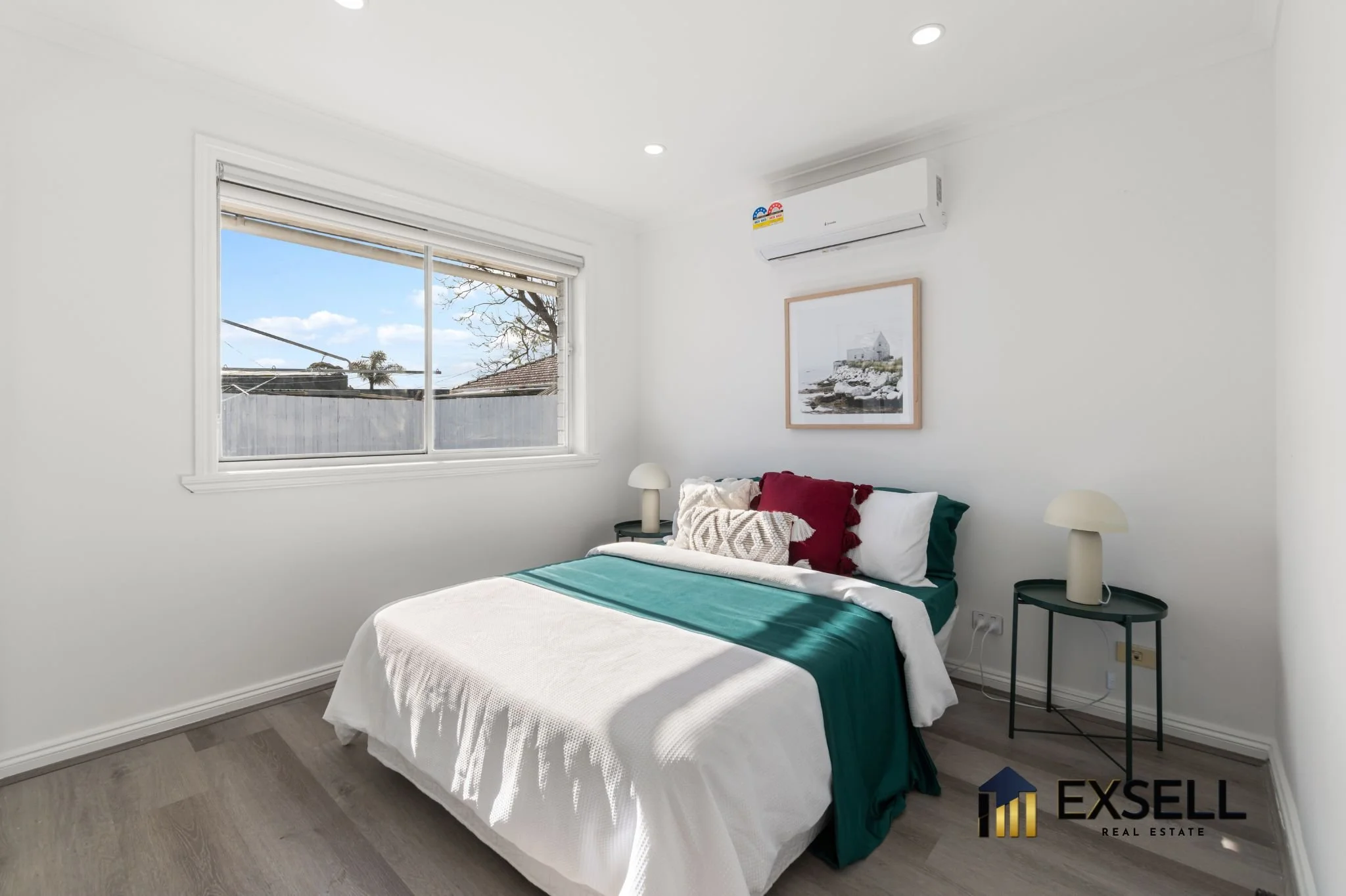 A bedroom with white walls, a window showing a tree and roofs outside, a double bed with white, green, and burgundy bedding and multiple pillows, two black side tables with white mushroom-shaped lamps, a framed landscape picture on the wall, and an a