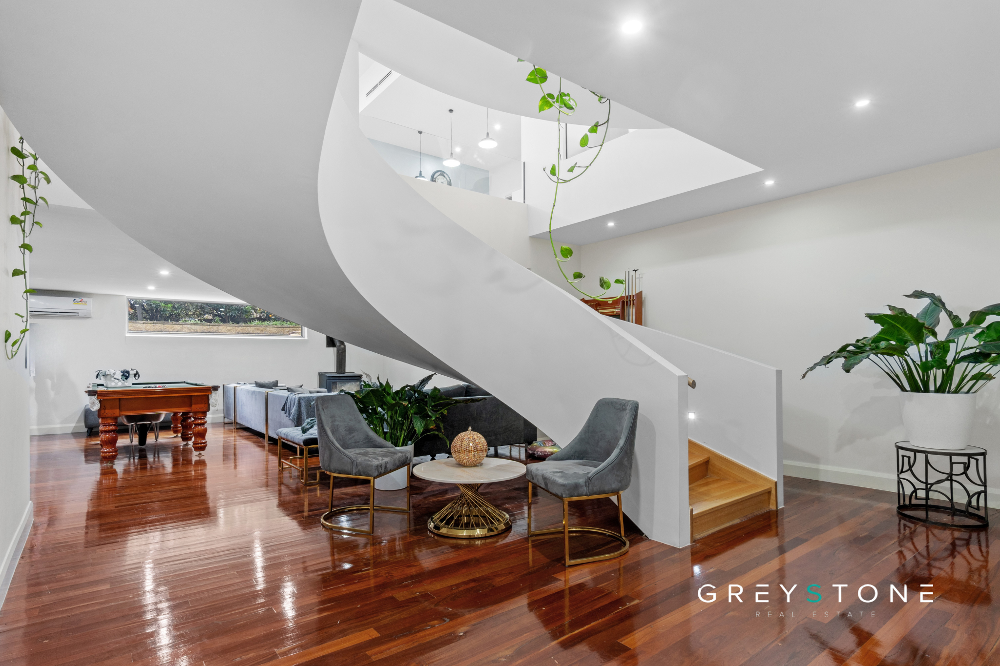 Modern living space with wooden floors, gray chairs around a small table, green plants, staircase, and a foosball table in the background.