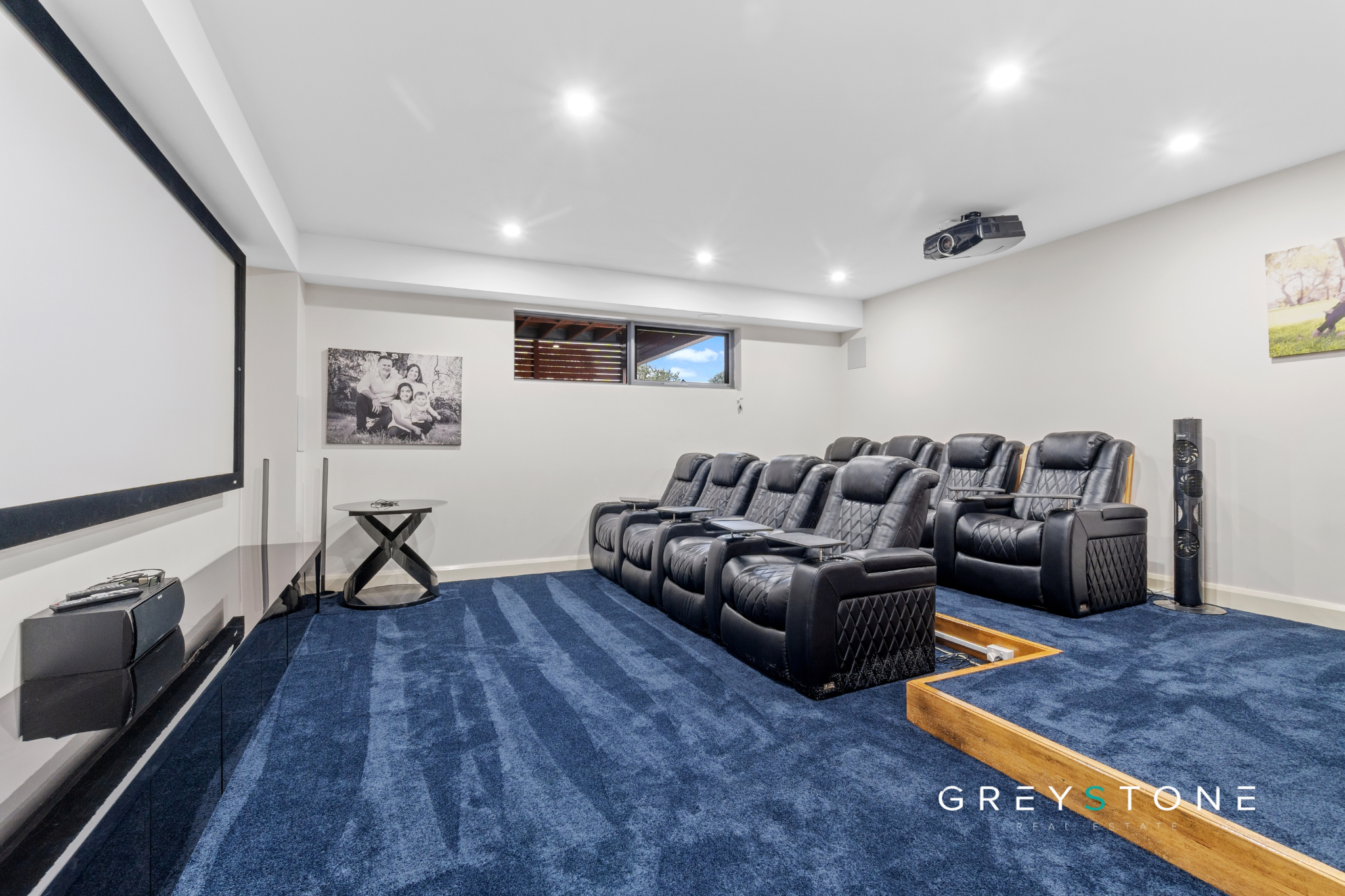 Home theater room with black leather recliners, a large projection screen on the wall, a ceiling-mounted projector, a window, family photos on the wall, and blue carpeting.