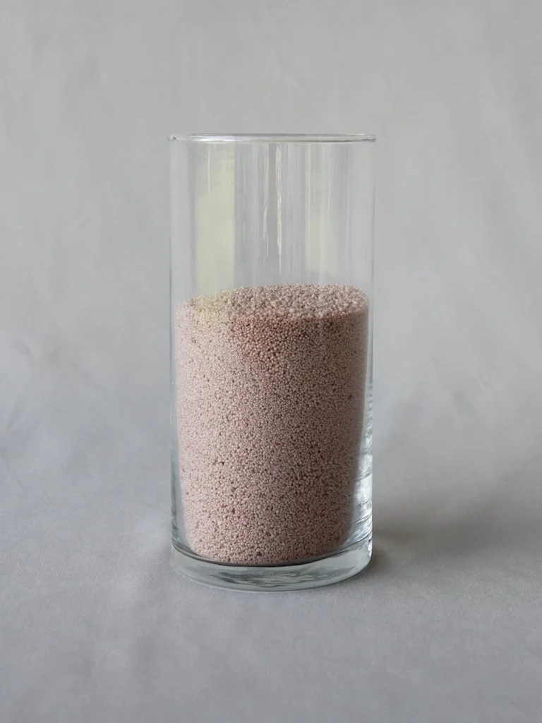 A clear glass filled with small, round, pinkish-grey granules against a plain white background.